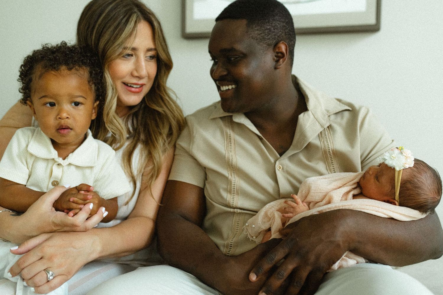 Happy interracial family with newborn baby and toddler sitting together on white couch smiling.