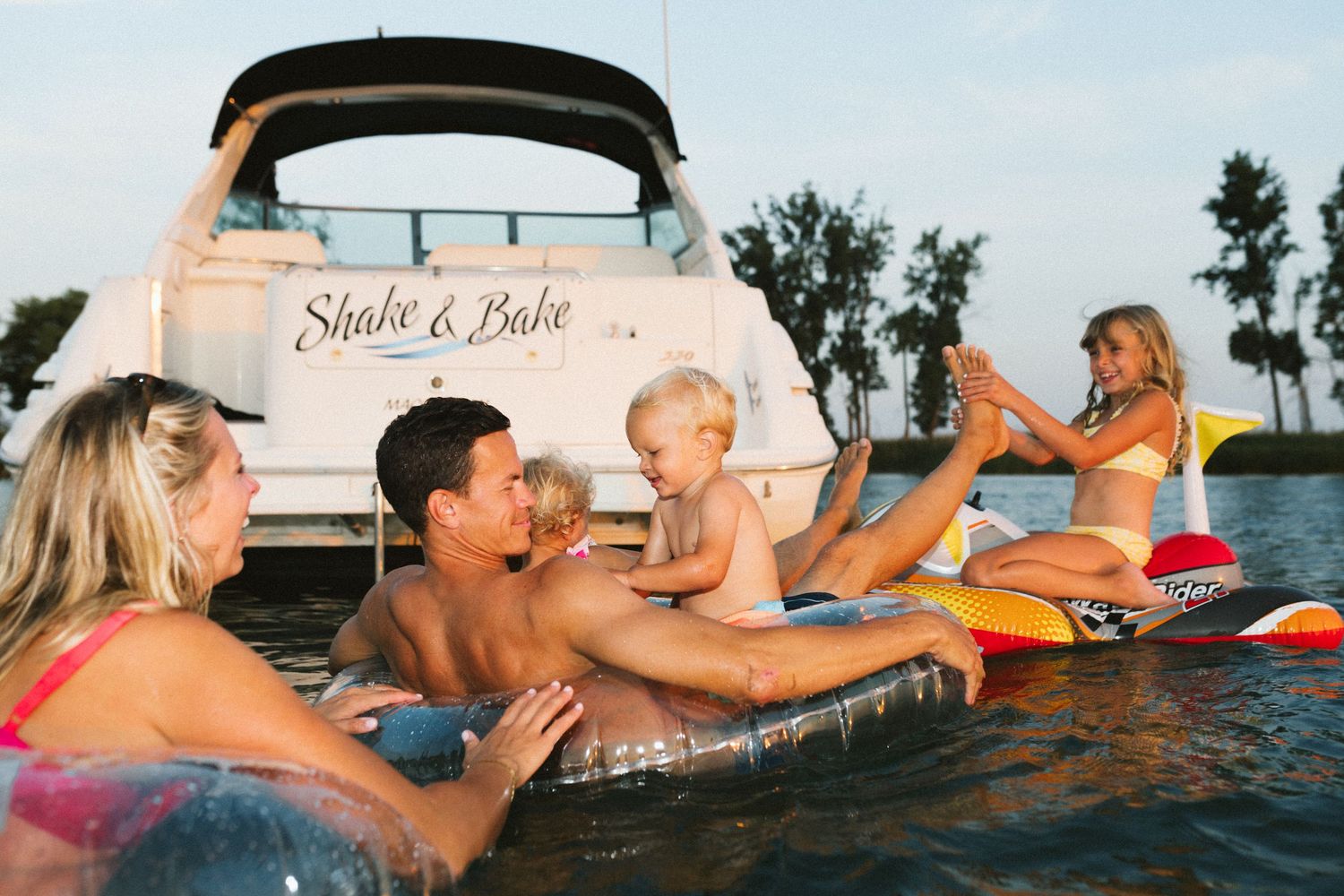 Family relaxing on inflatable floats beside a boat named Shake & Bake on a calm lake at sunset.