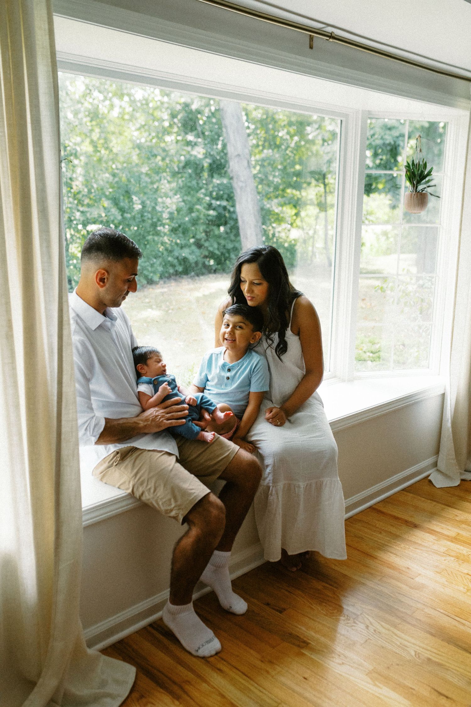 Family of four sitting by large window with newborn baby in a bright sunlit living room.