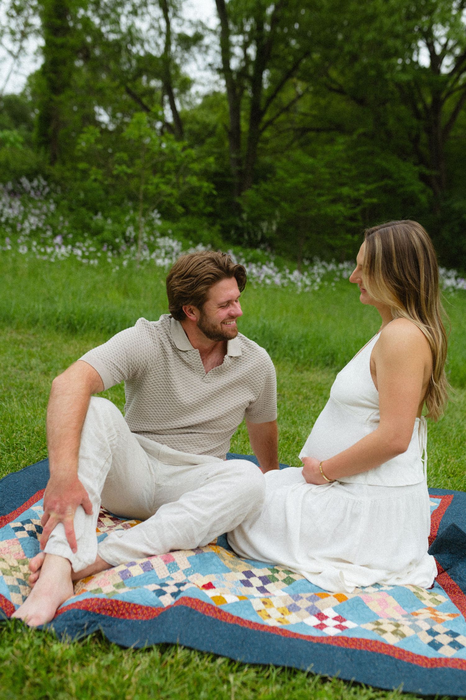 Happy couple sitting on patchwork quilt in green park during maternity photo session.