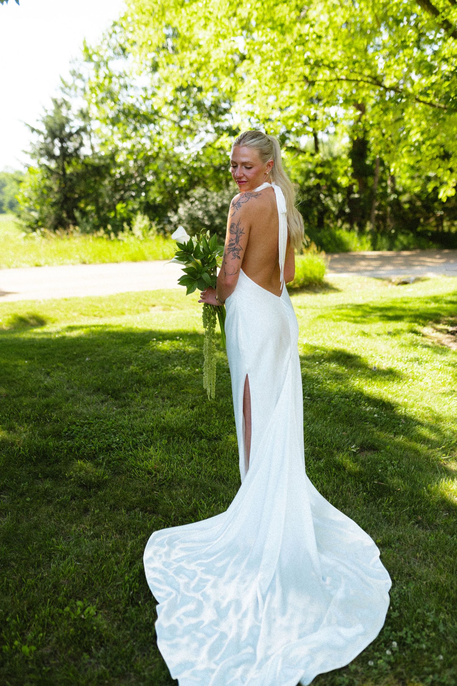 Bride in backless white gown with train holds bouquet outdoors.