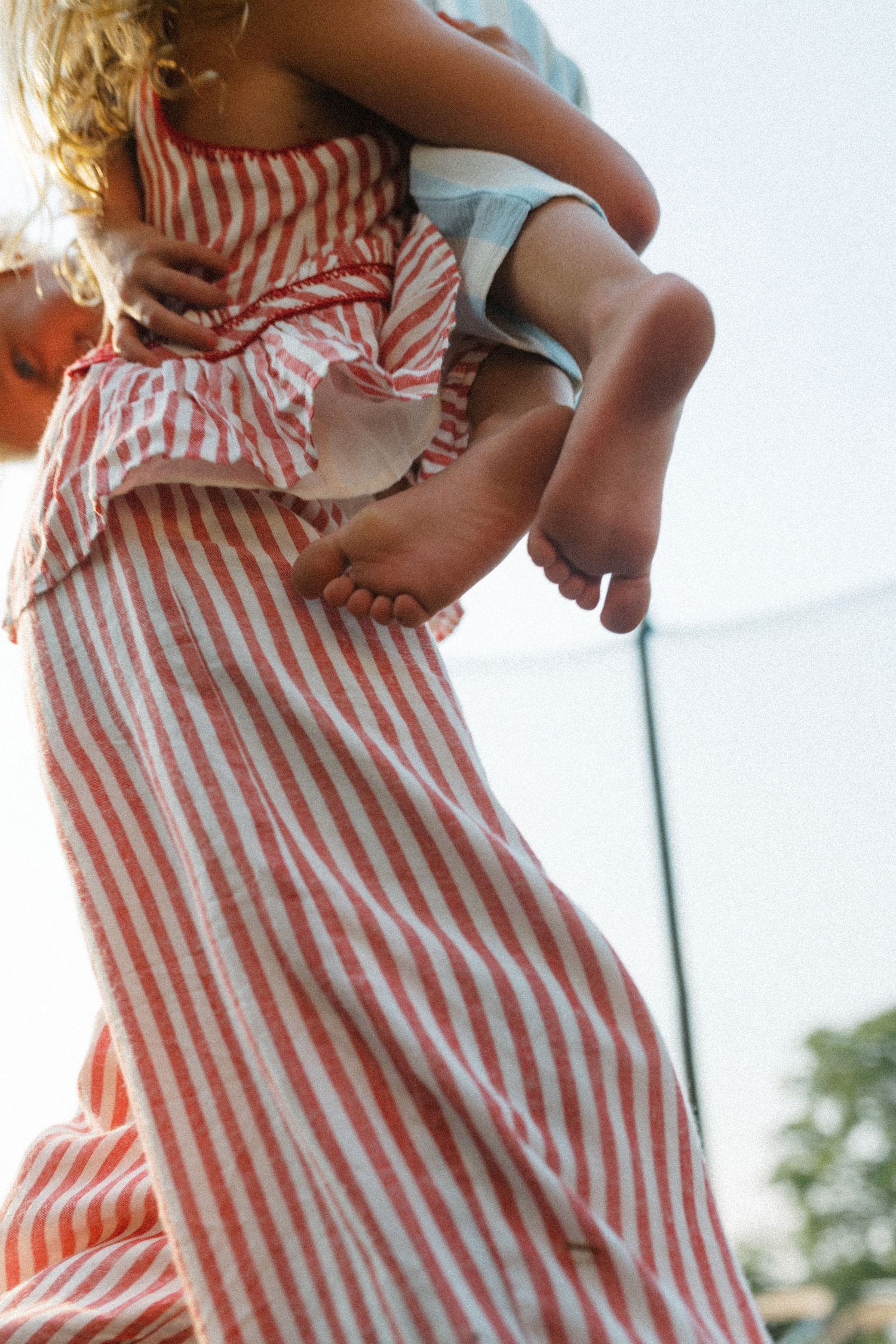 Woman in red striped dress carrying a young child barefoot on a sunny outdoor trampoline.