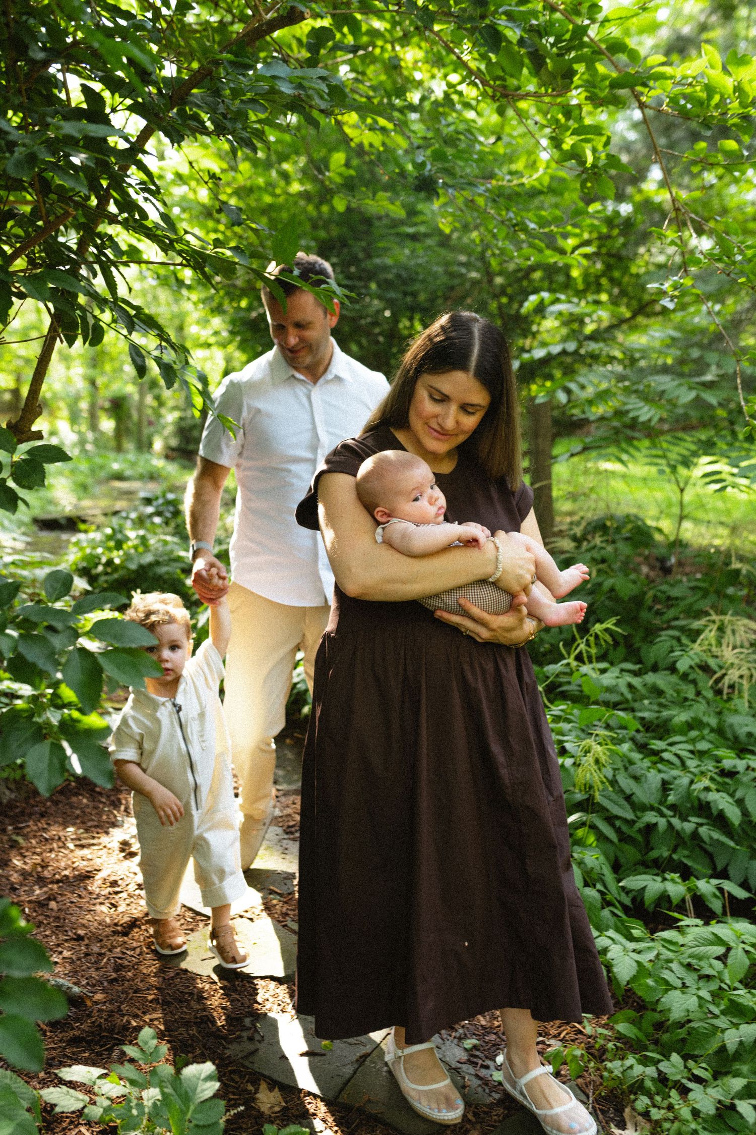 Young family walking through sunlit green forest path, mother cradling newborn in dark dress, father and toddler following.