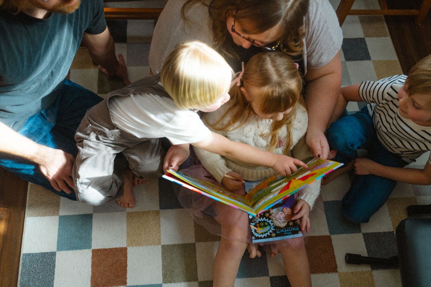Family sitting together on a checkered floor reading a colorful picture book.