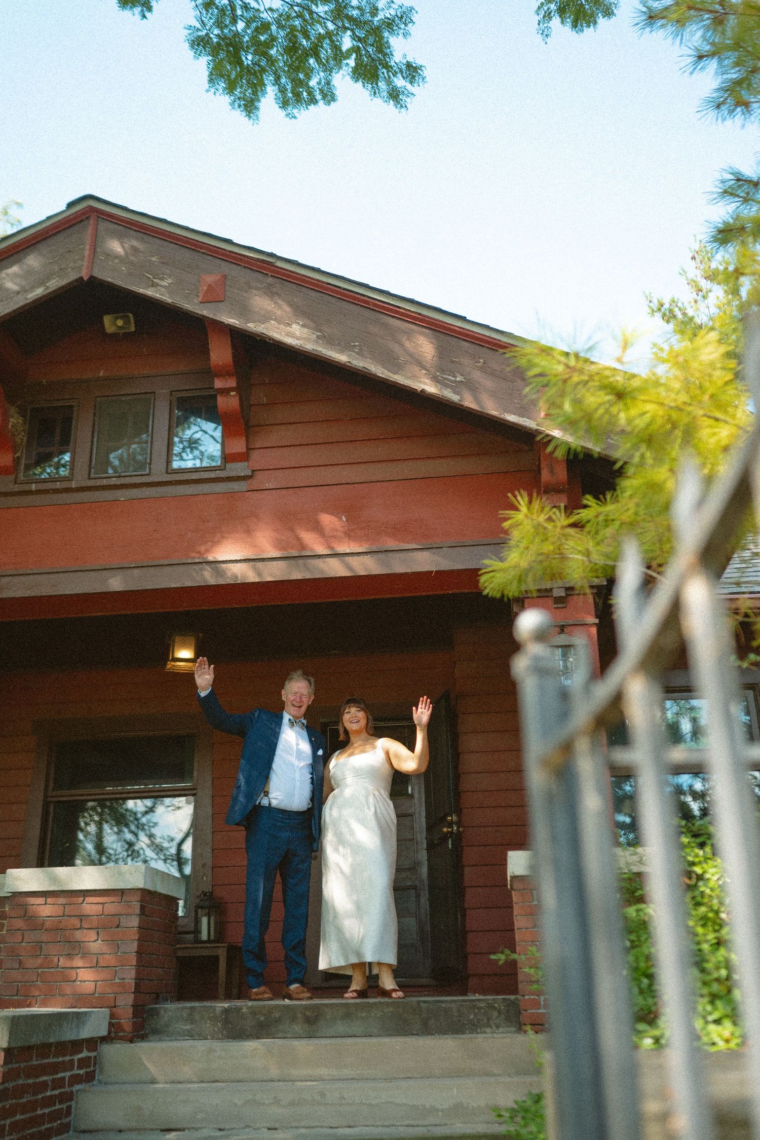 Couple in wedding attire waving from the porch of a charming red craftsman house on their wedding day.