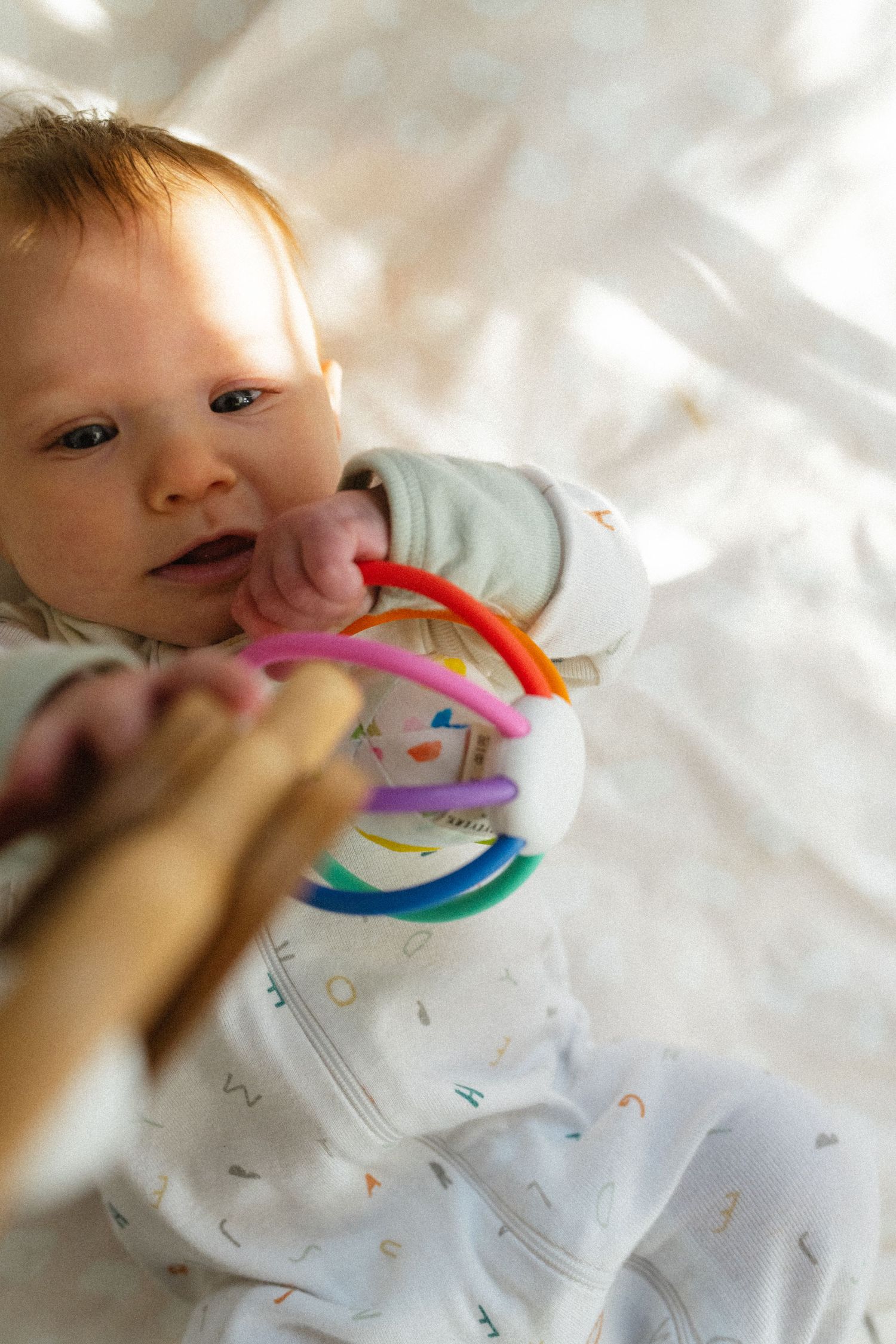 Newborn baby in white onesie chewing on colorful ring toy while lying on a soft white blanket.