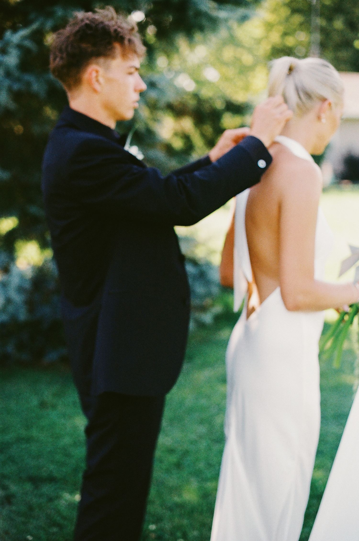 Groom fastening the back of a brides white wedding dress outdoors in garden.