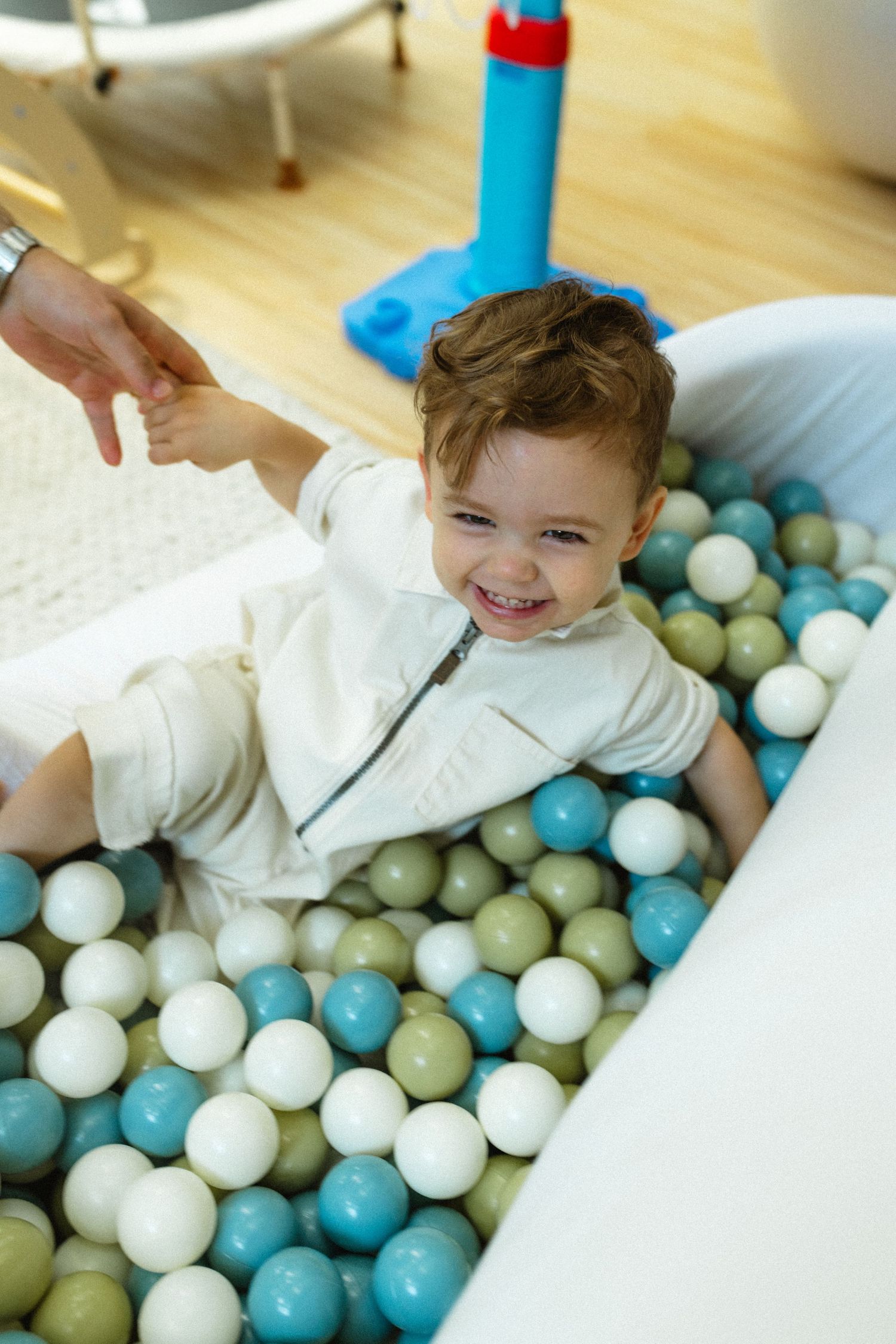 Happy toddler boy smiling in colorful ball pit while adult reaches out hand to help.