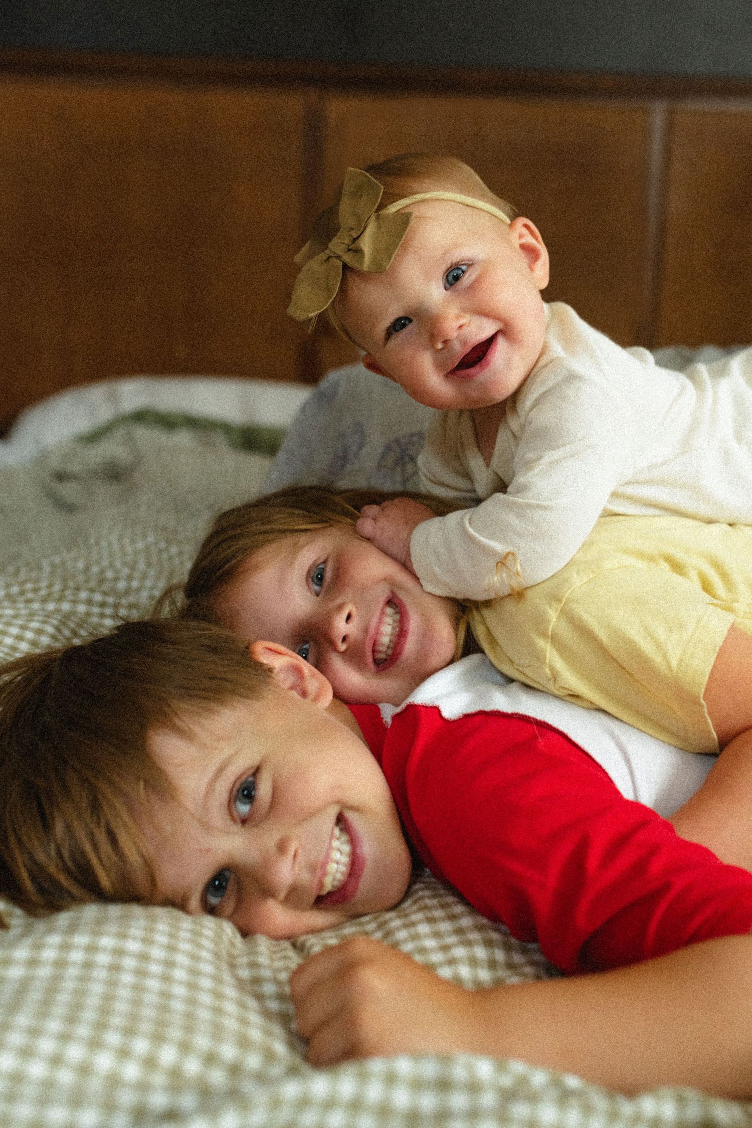 Three smiling siblings lying on a bed, with the youngest baby girl wearing a yellow bow headband on top.