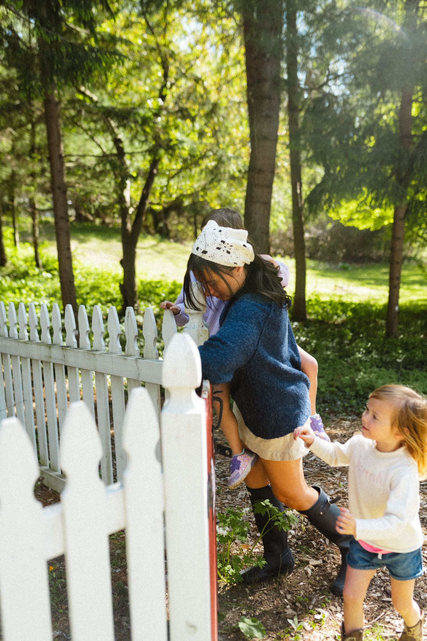 Woman lifting a child over a white picket fence gate in a sunny green garden setting.