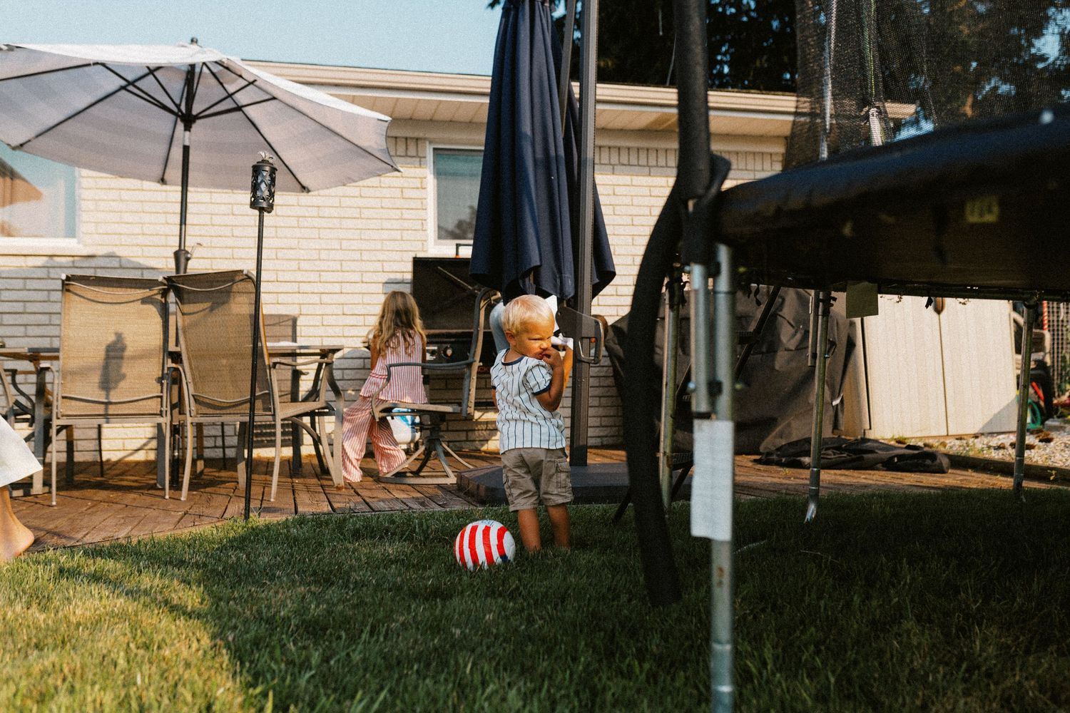 Two young children playing in a sunny backyard near a trampoline and patio.