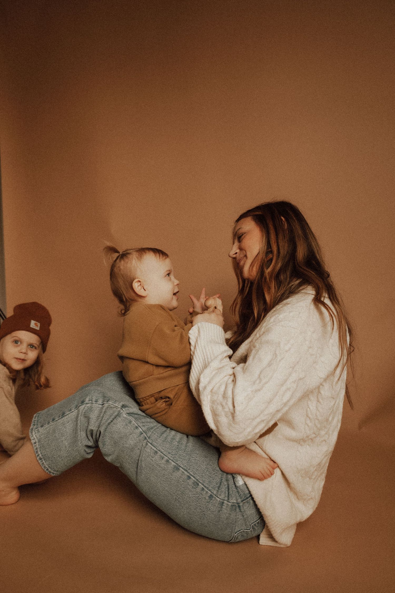 A mother sitting on the floor smiling and playing with her toddler while another child watches nearby.