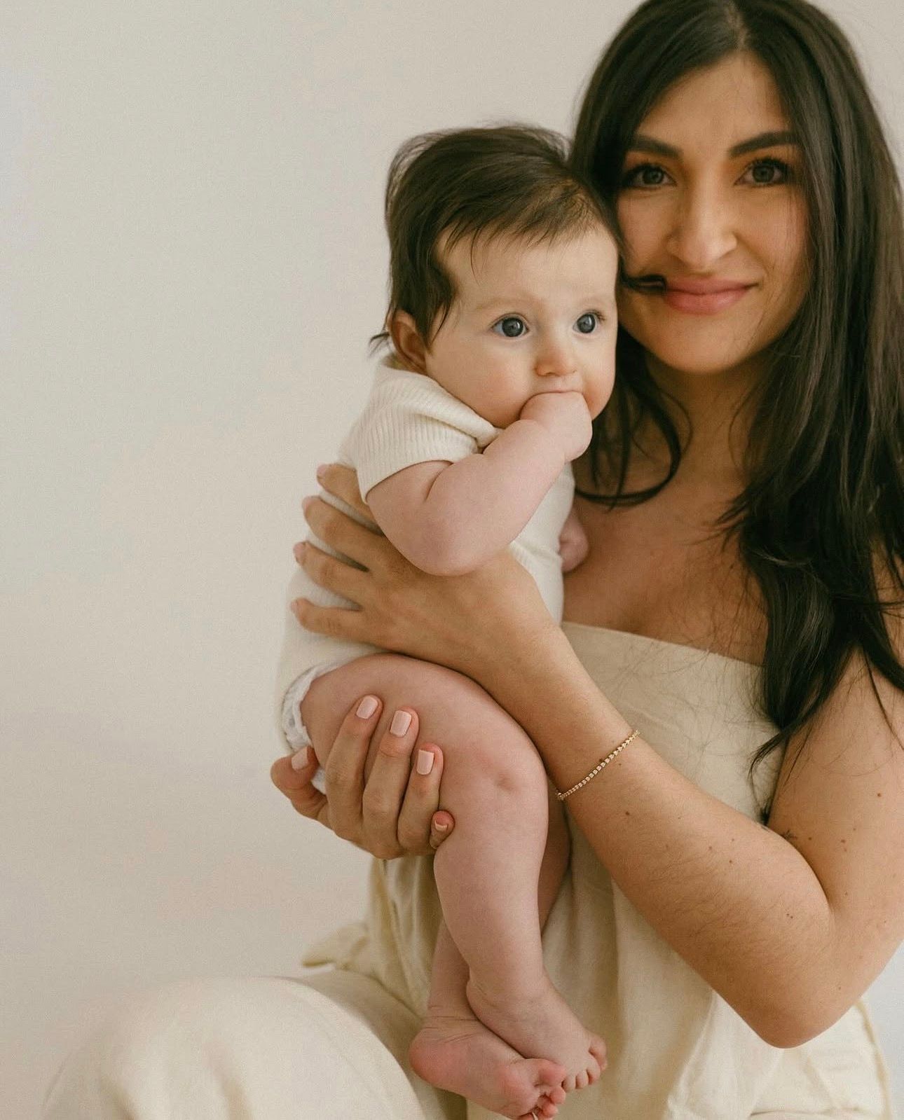 A smiling brunette woman in a white dress holds an adorable baby in a white onesie against a light background.