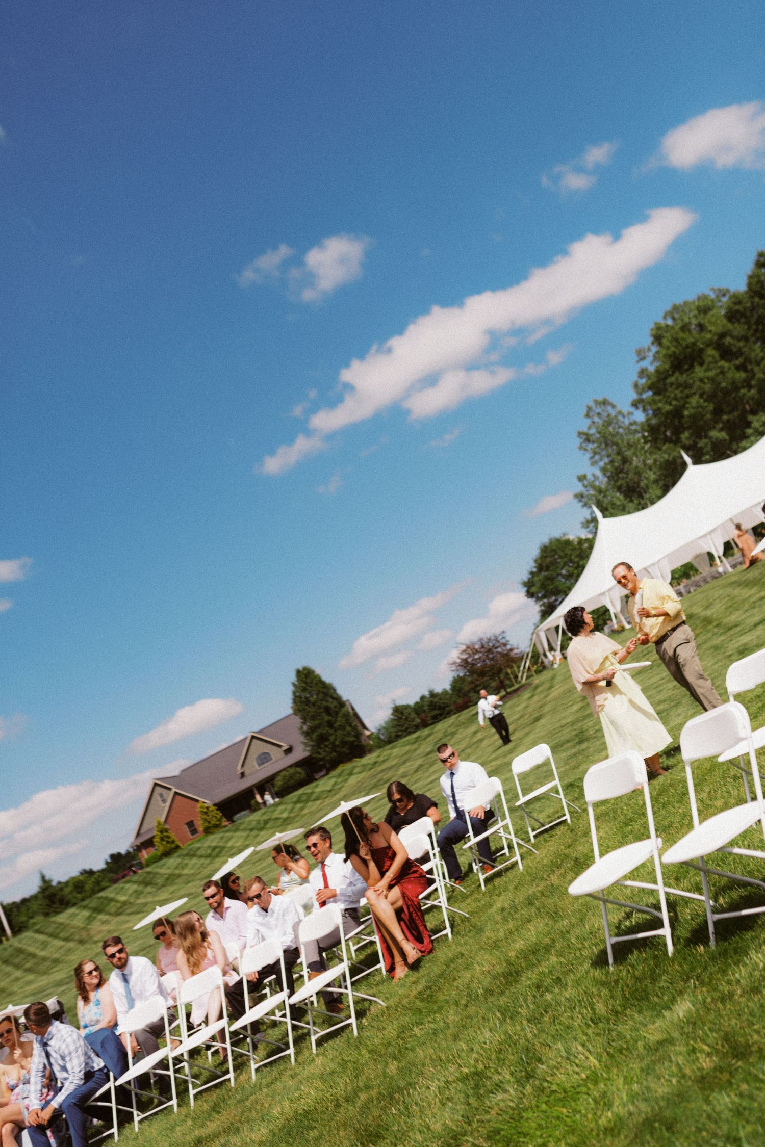 Outdoor wedding ceremony with guests seated in white chairs on sunny green lawn.