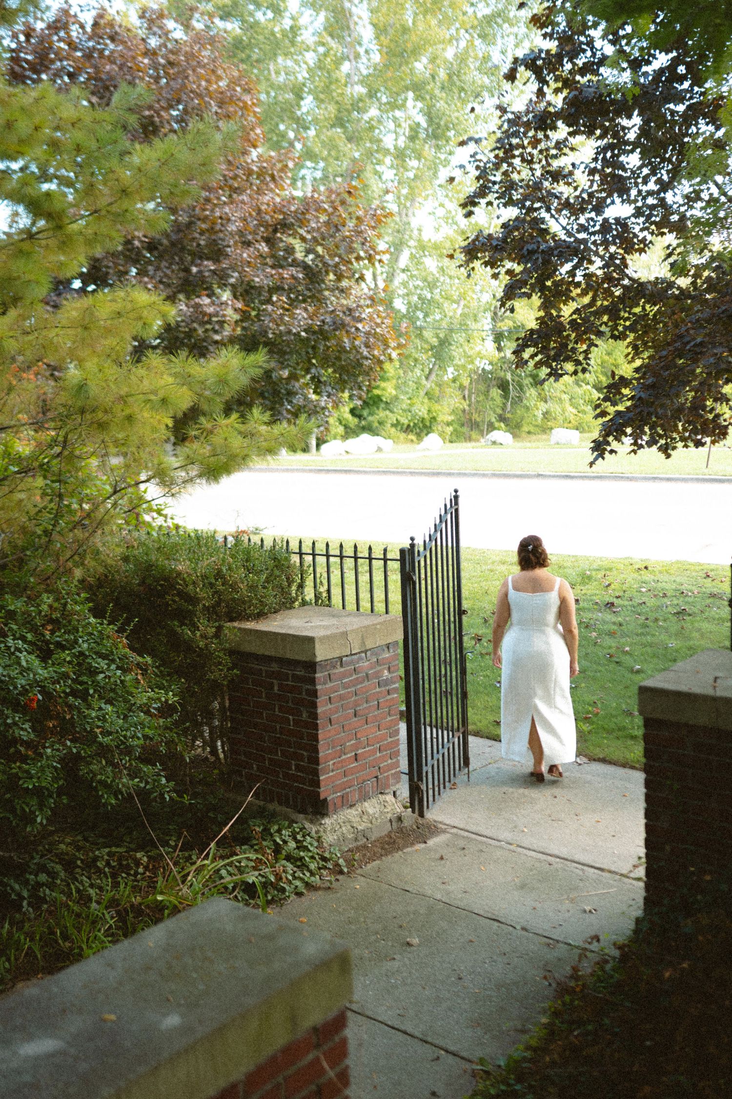 Bride in white gown walking through iron garden gate surrounded by lush green trees.