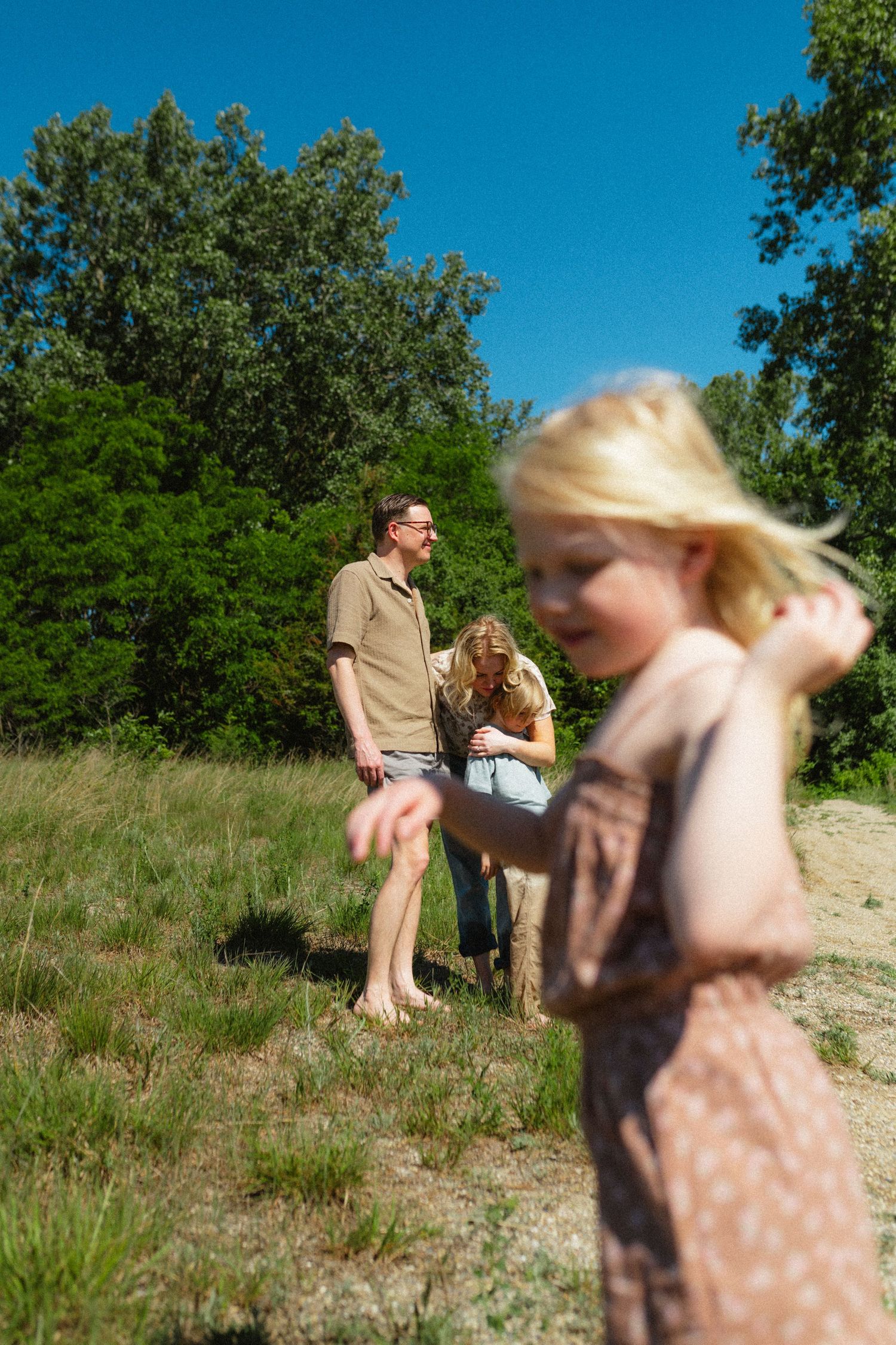 Blonde girl in foreground with family standing on grassy hill surrounded by green trees on sunny day.