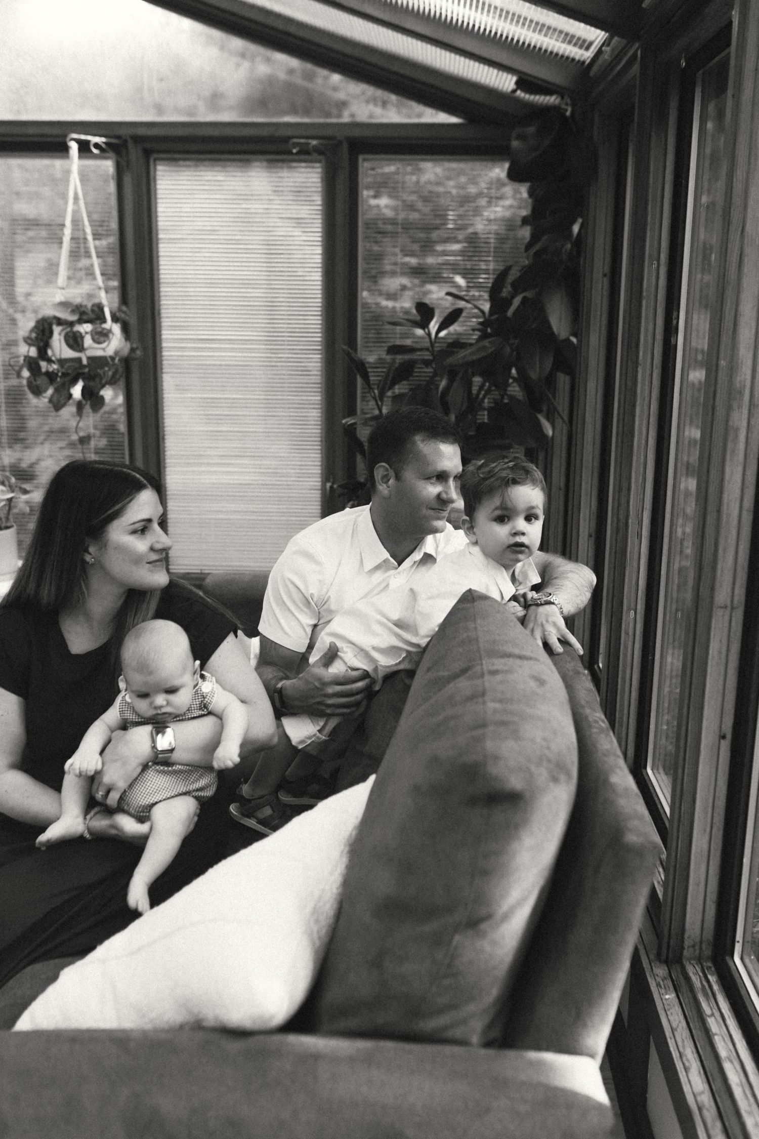 Black and white photo of family with baby and toddler relaxing together on sofa in sunroom.