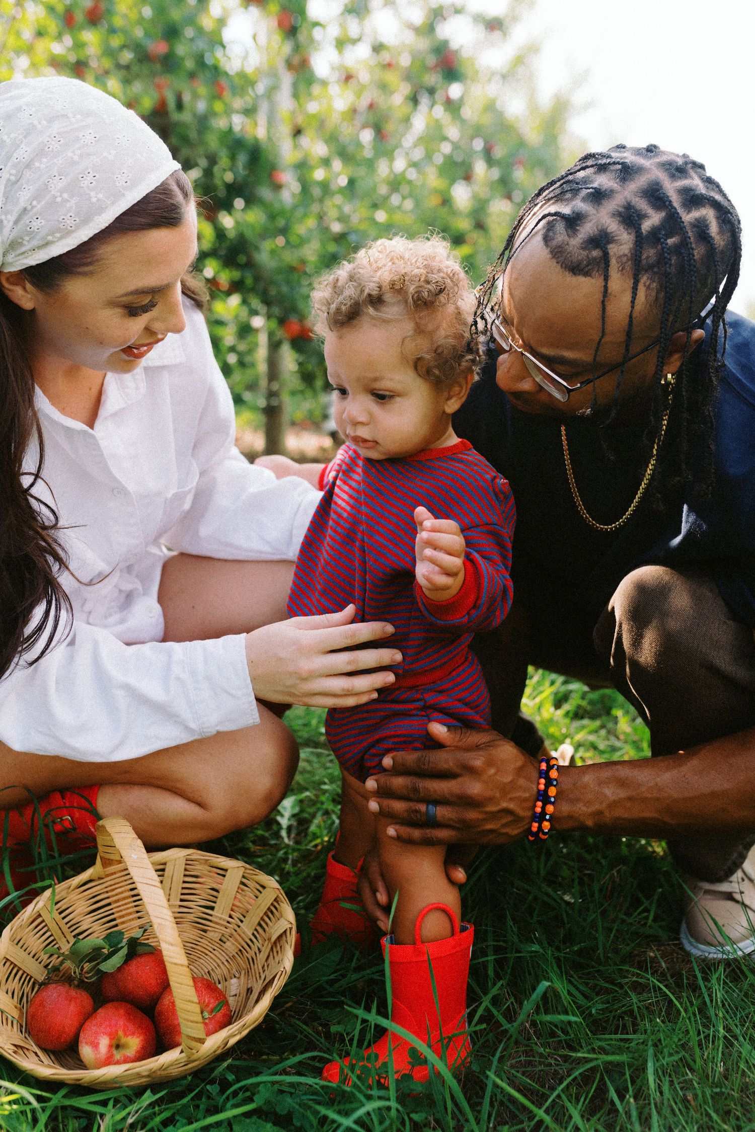 A diverse family picks apples together outdoors, with a toddler in red boots standing by a wicker basket full of apples.