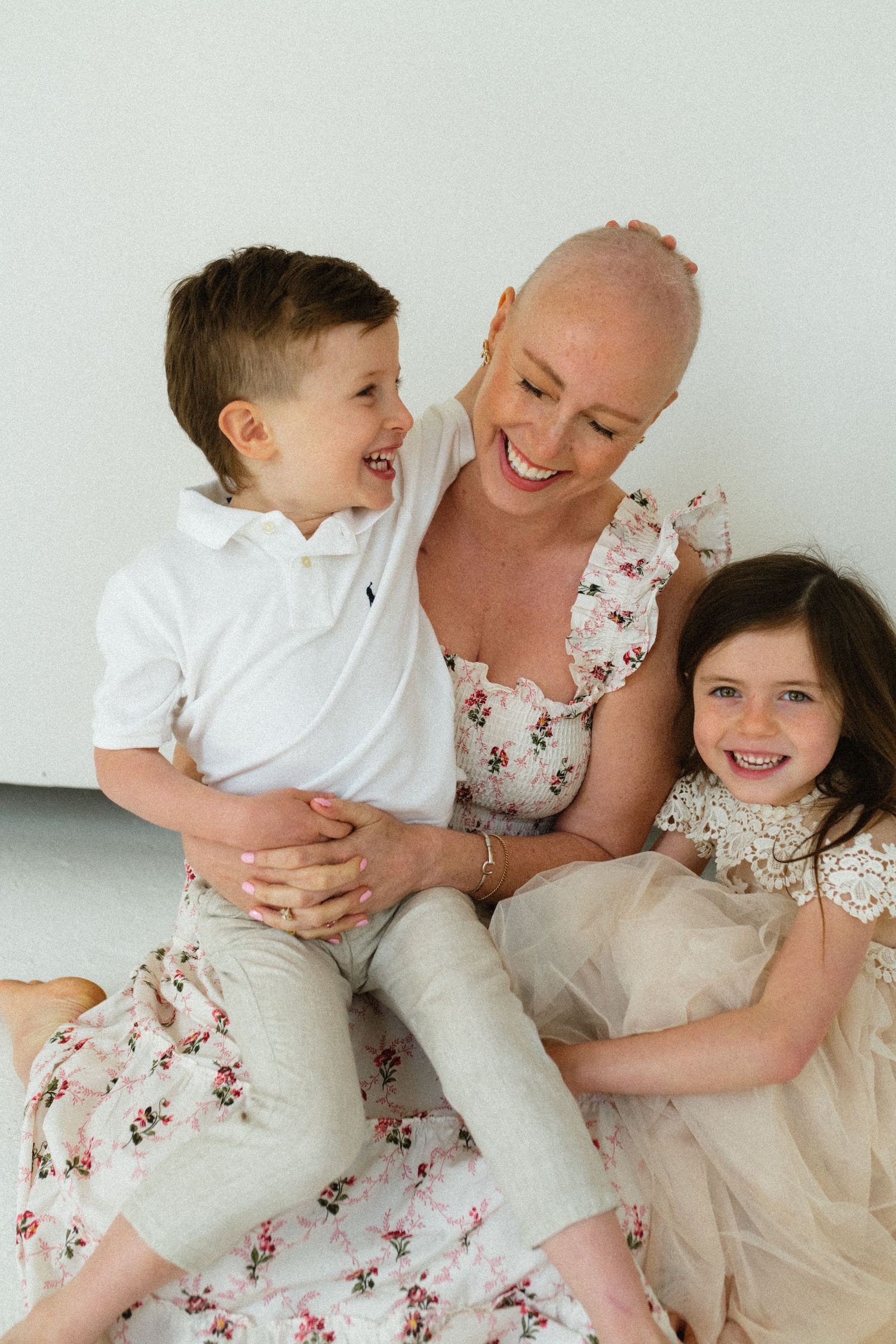 Bald woman smiling and laughing with two young children in a bright white studio.