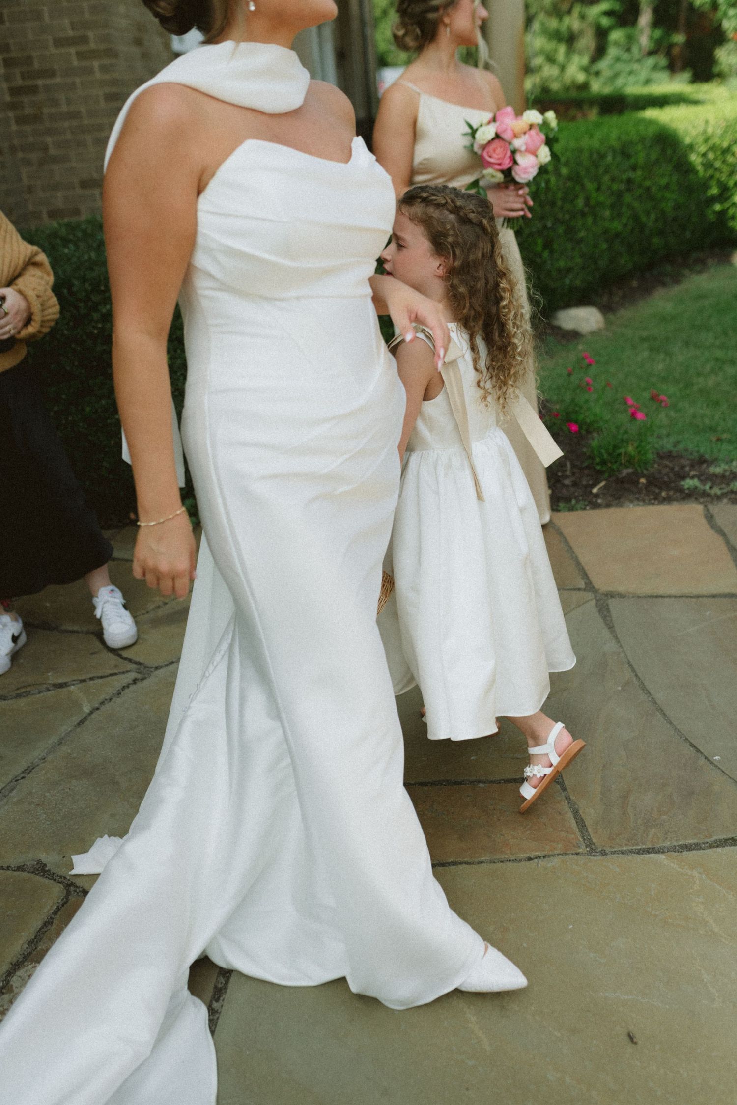 Flower girl in white dress hugging bride in elegant halter wedding gown on stone patio.