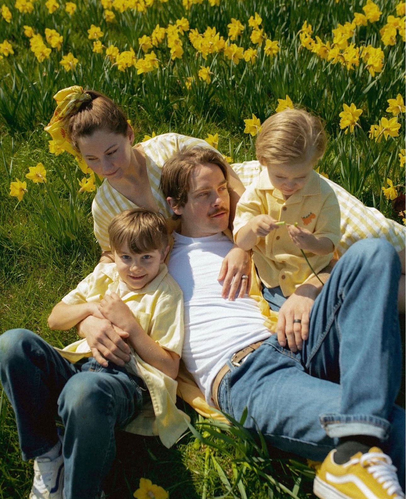 Family of four relaxing in a sunny daffodil field, wearing coordinating yellow outfits on a bright spring day.