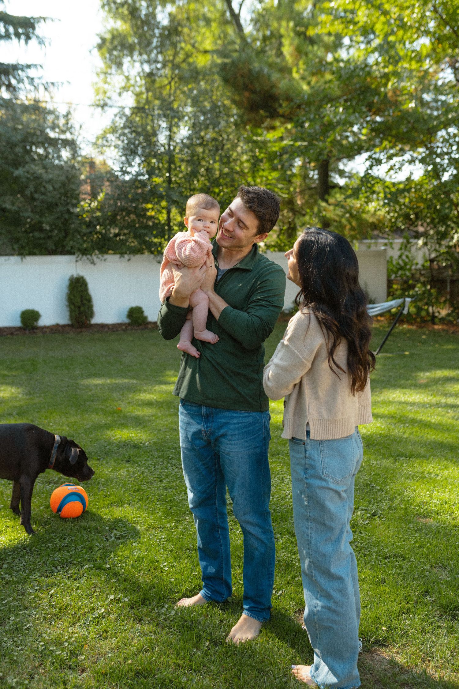 Smiling couple holding their baby outdoors in a sunny backyard with a black dog playing nearby.