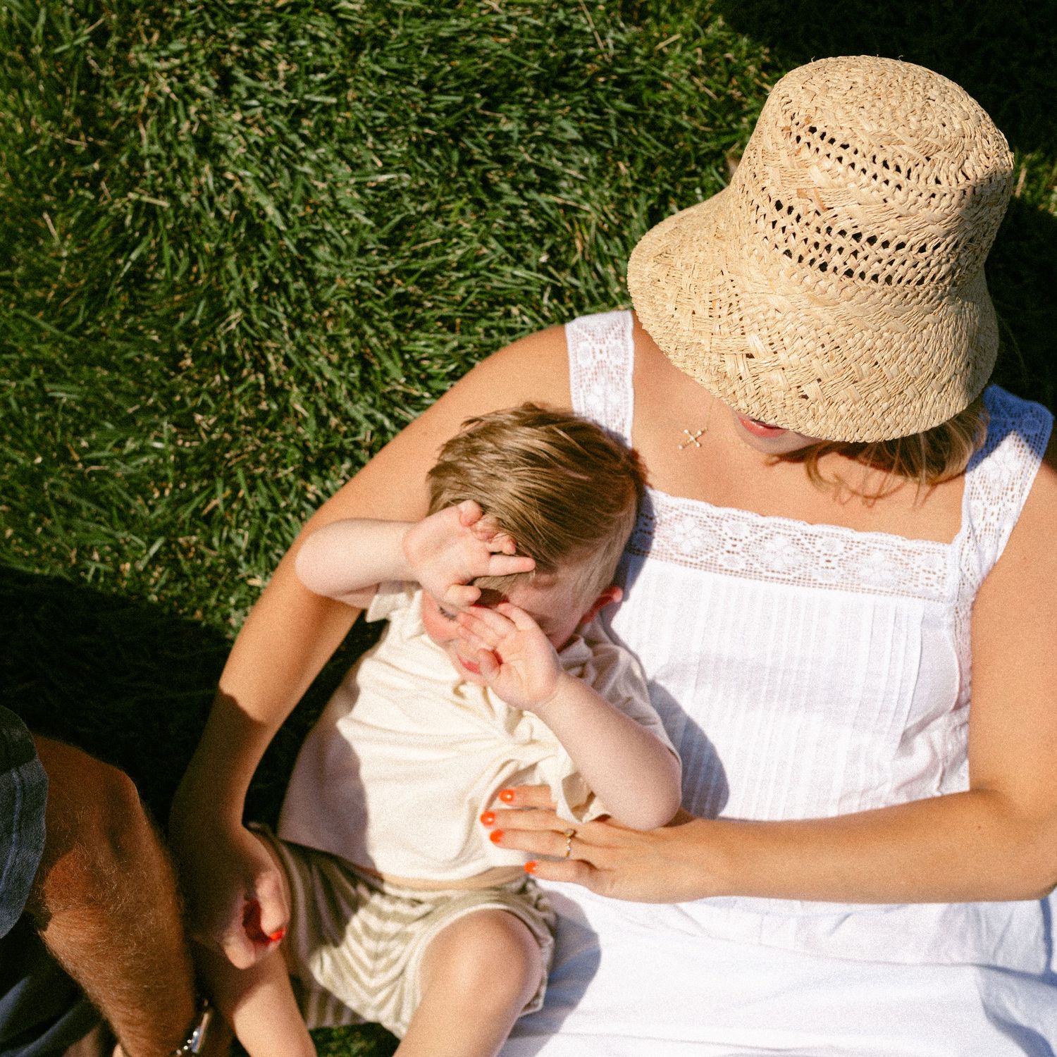 Mother in straw hat and white dress cradling young child on green grass in bright sunlight.