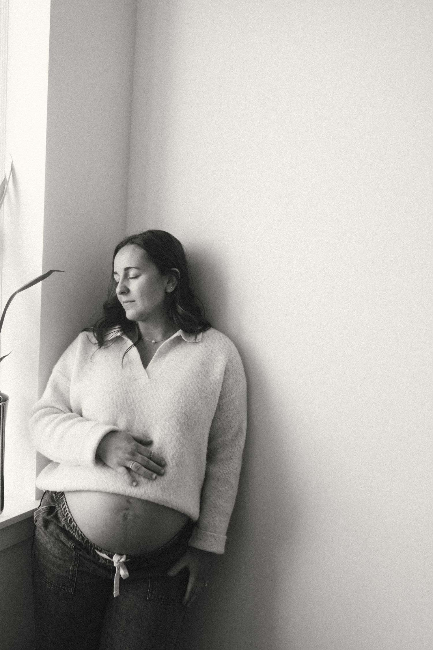 Black and white portrait of a pregnant woman leaning against a wall with eyes closed, cradling her belly.