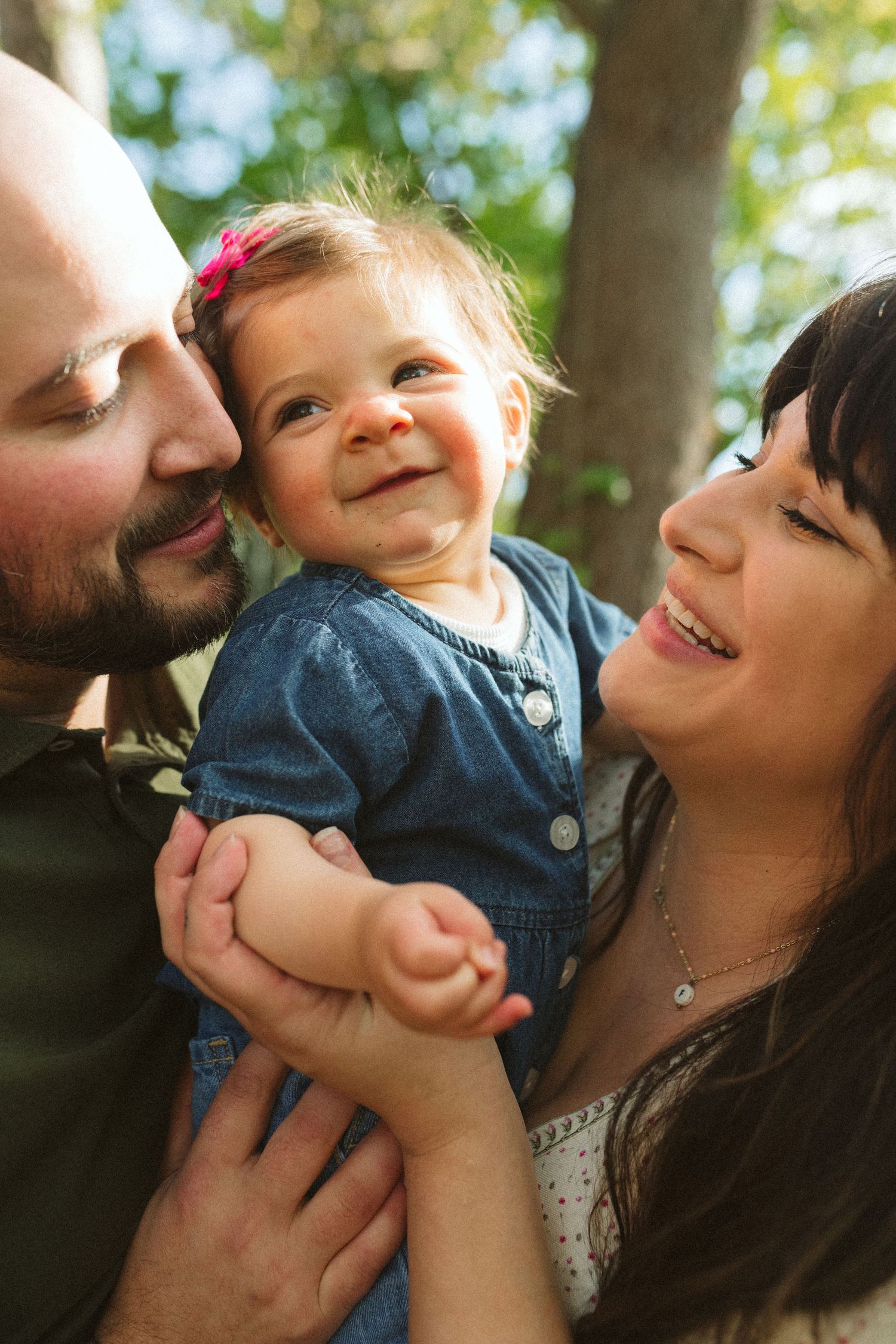 Smiling parents lovingly holding their happy baby girl outdoors among green trees.