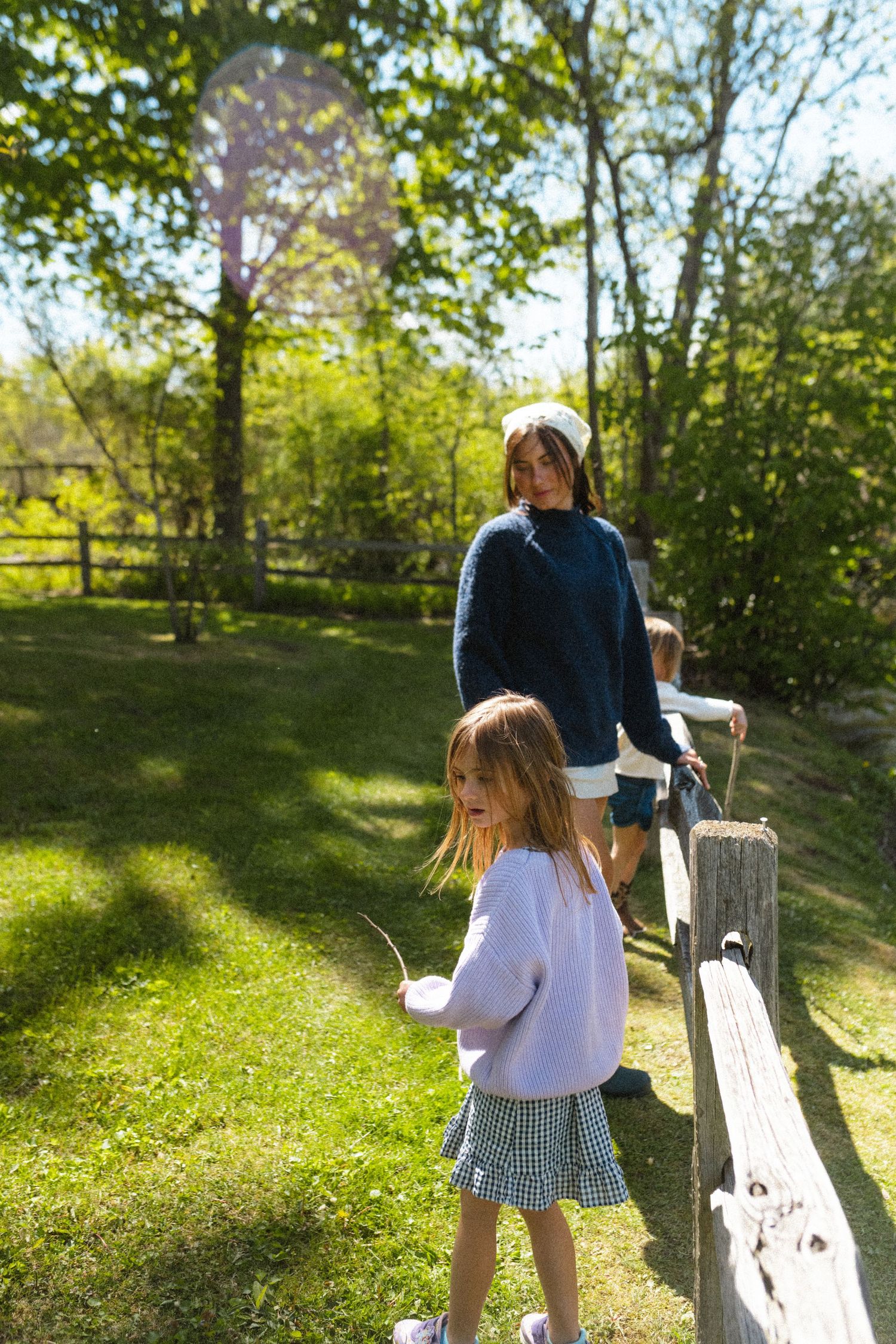 Mother and two young daughters walking along a wooden fence in a sunny green garden.