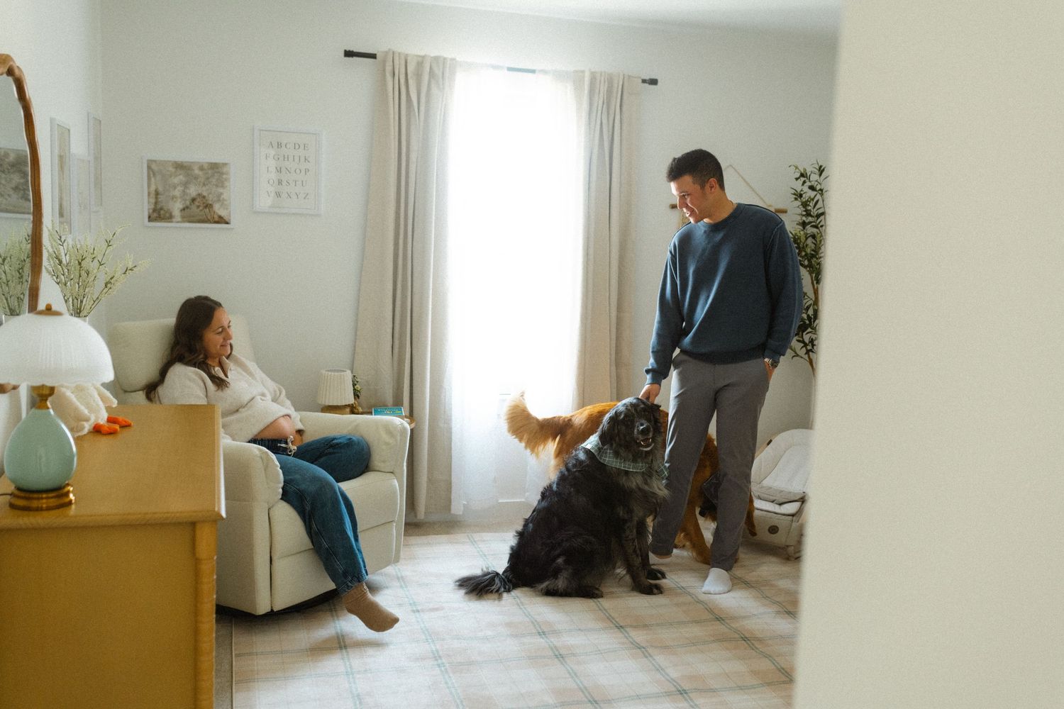 Couple playing with large fluffy dog in bright living room, woman relaxing on sofa watching man interact with pet.