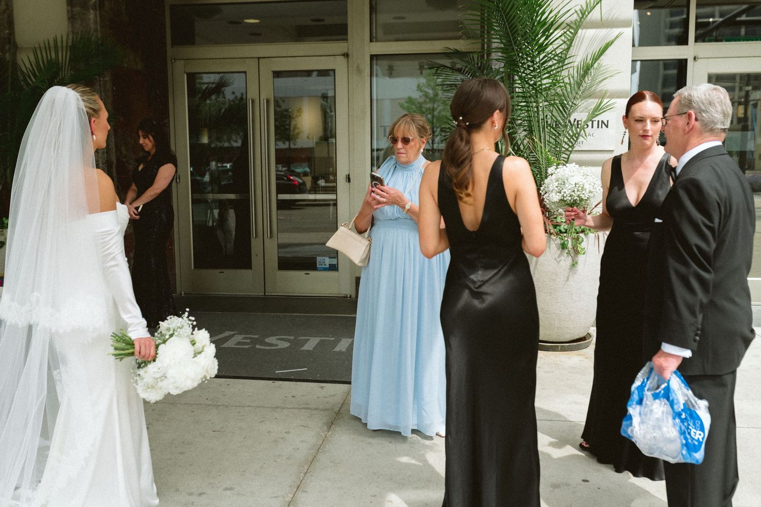Bride in white veil greets bridesmaids in black and blue gowns outside urban wedding venue.