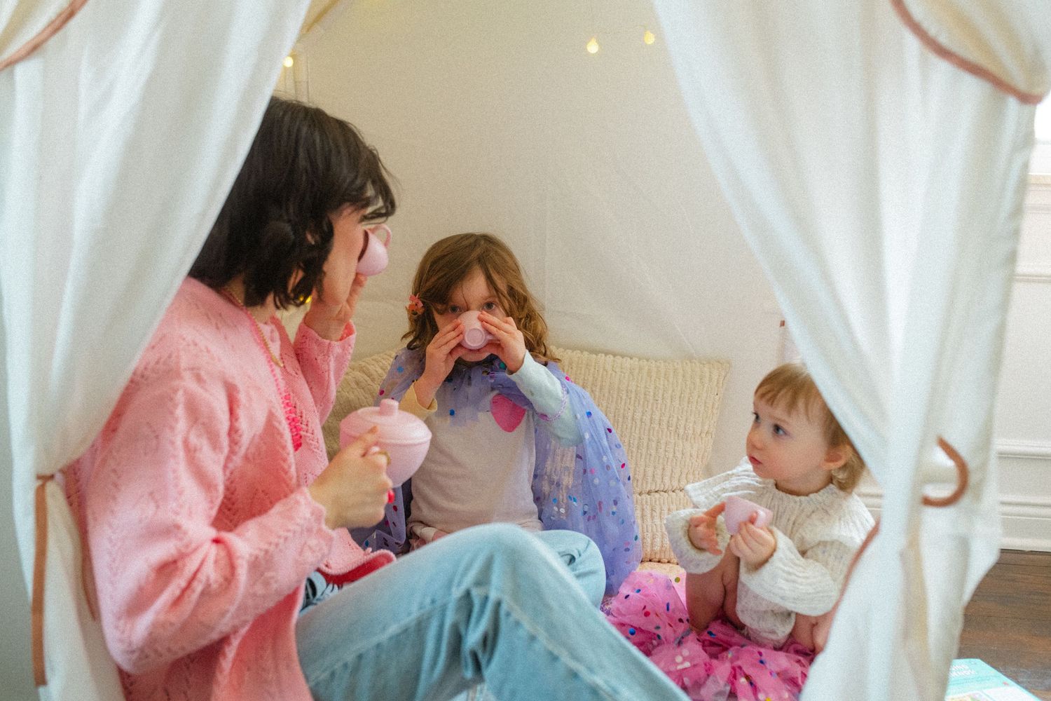 Mother and two young children playing together inside a white fabric play tent with fairy lights.