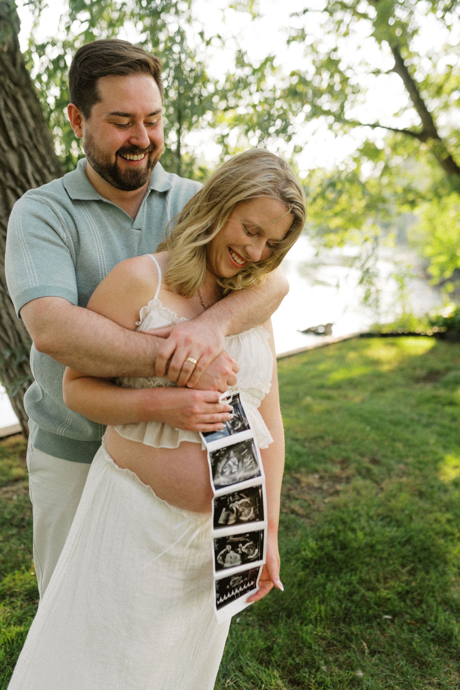 A smiling couple holding ultrasound photos while embracing outdoors near a lake during golden hour.