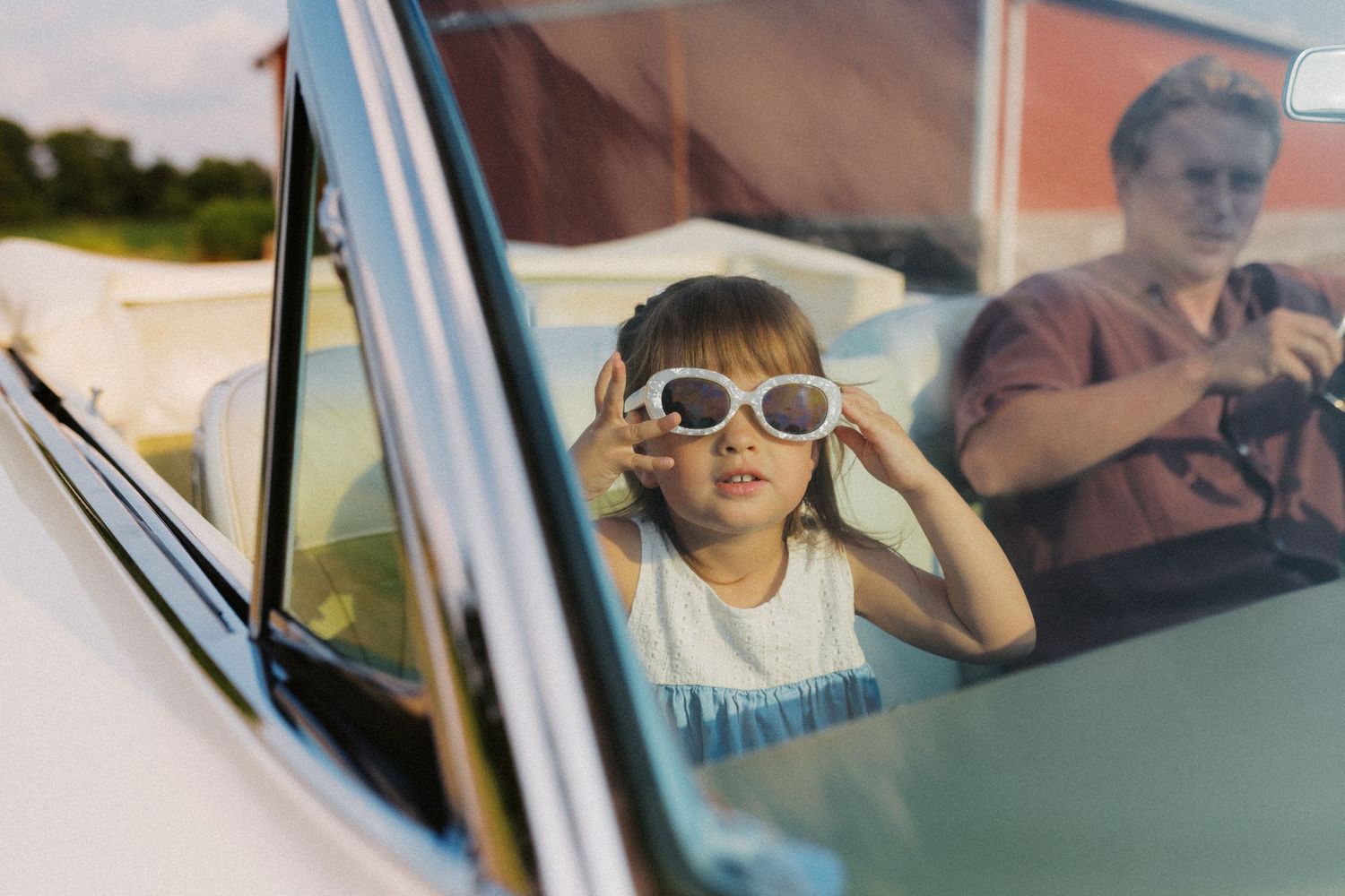 Young girl trying on white sunglasses inside a classic vintage convertible car.
