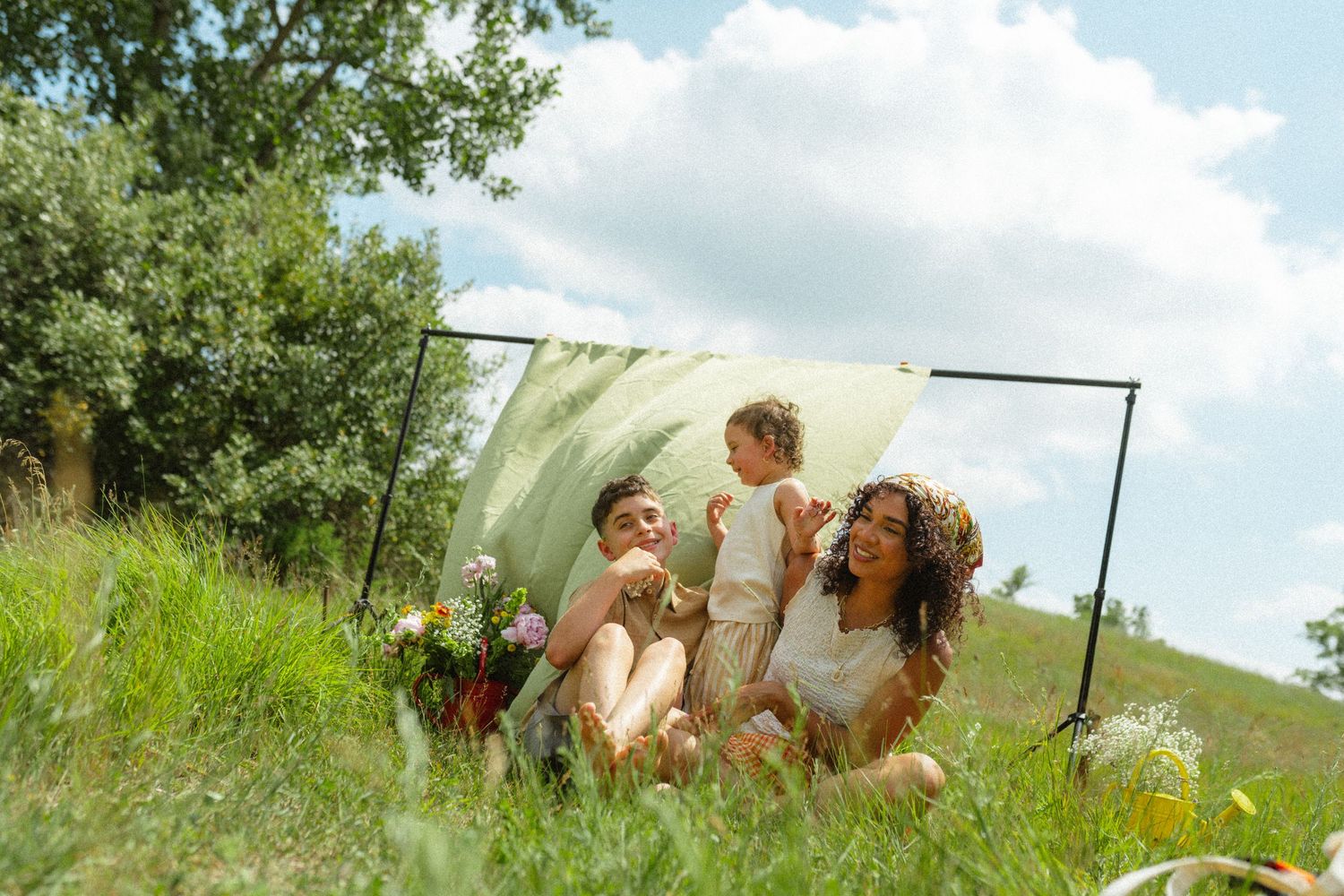 Mother and two children laughing together on green grass with a fabric backdrop outdoors.