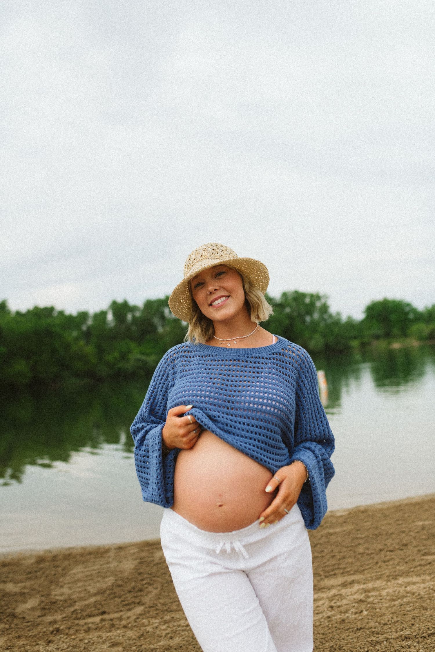 Smiling pregnant woman in blue knit sweater and straw hat holding baby bump by lakeside.