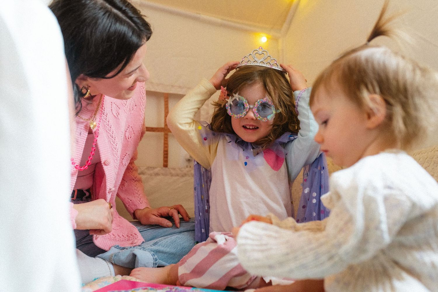 Young girl wearing princess crown and novelty sunglasses playing with mother and sibling in tent.