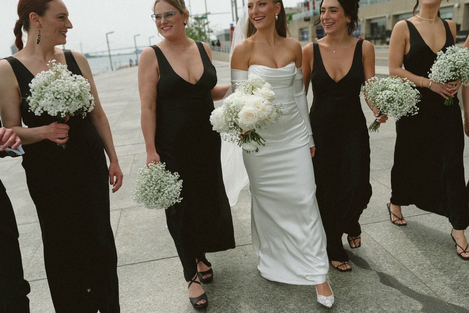 Bride in sleek white gown walks with bridesmaids in black dresses holding white floral bouquets.