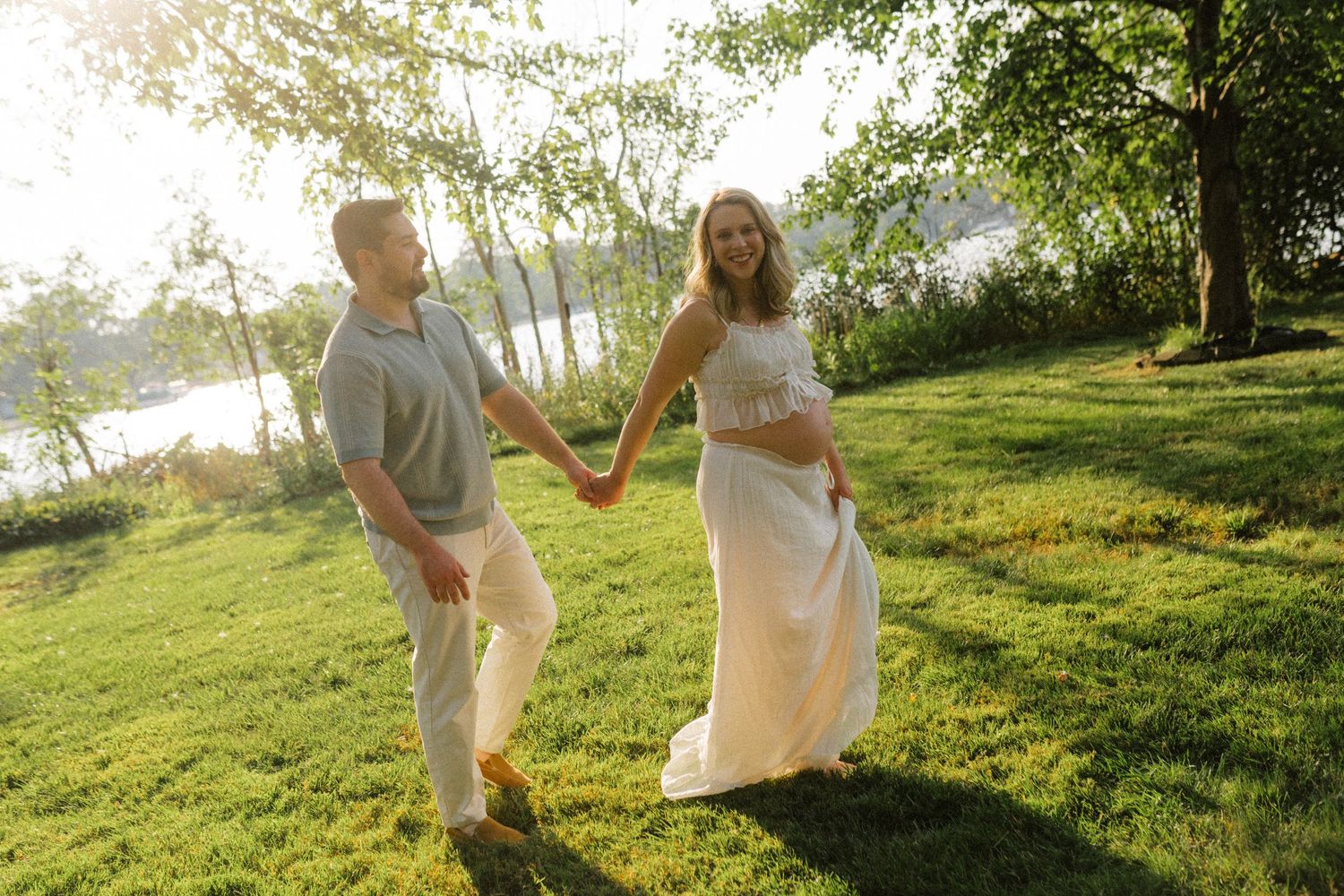 A happy couple holding hands and walking on green grass near a sunlit lake under a tree.