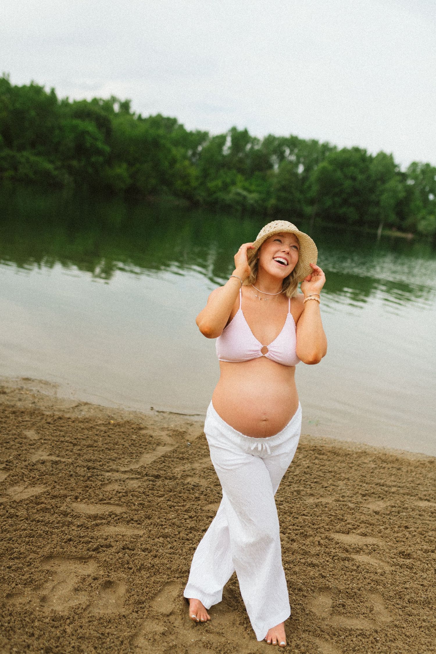 Joyful pregnant woman in pink bikini top and white pants adjusting straw hat on sandy beach.