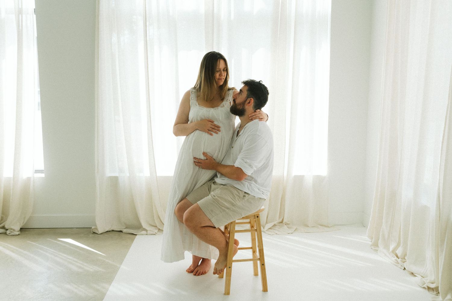 Pregnant woman in white dress with bearded partner in bright studio maternity photo session.