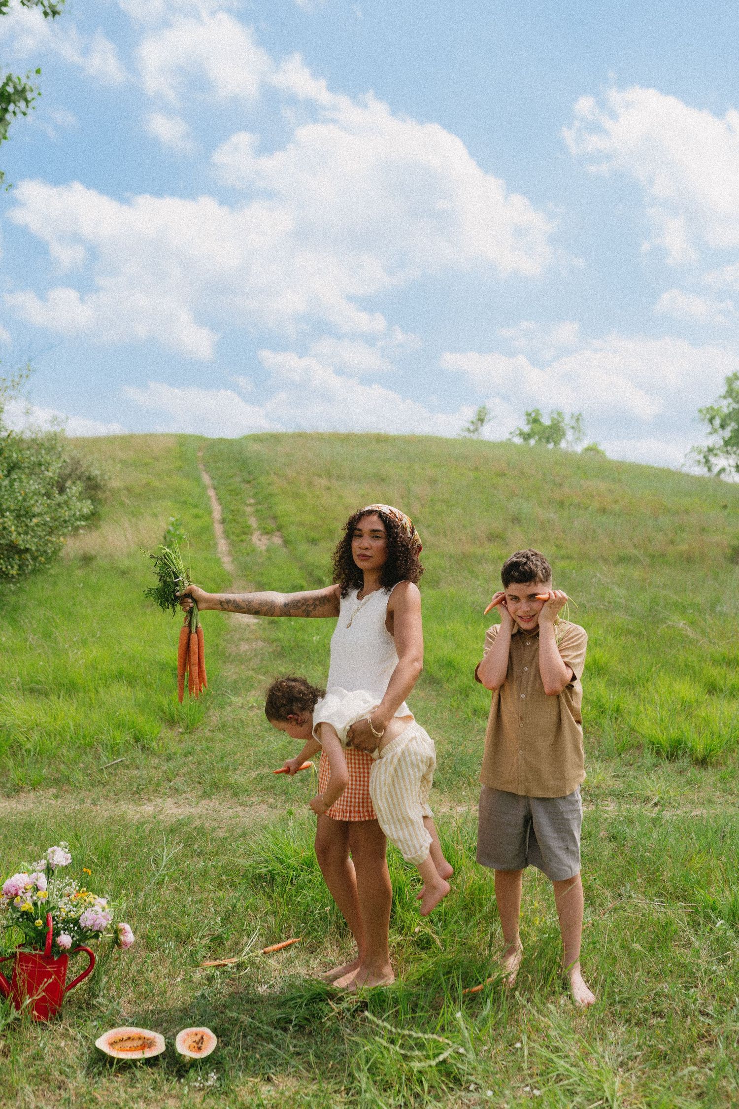 Mother holding toddler and carrots stands with son on a scenic green hillside outdoors.