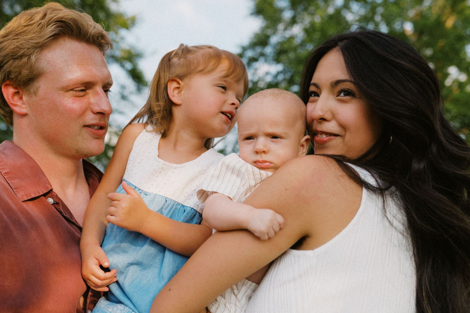 Close-up portrait of smiling parents holding two young children outdoors in park.