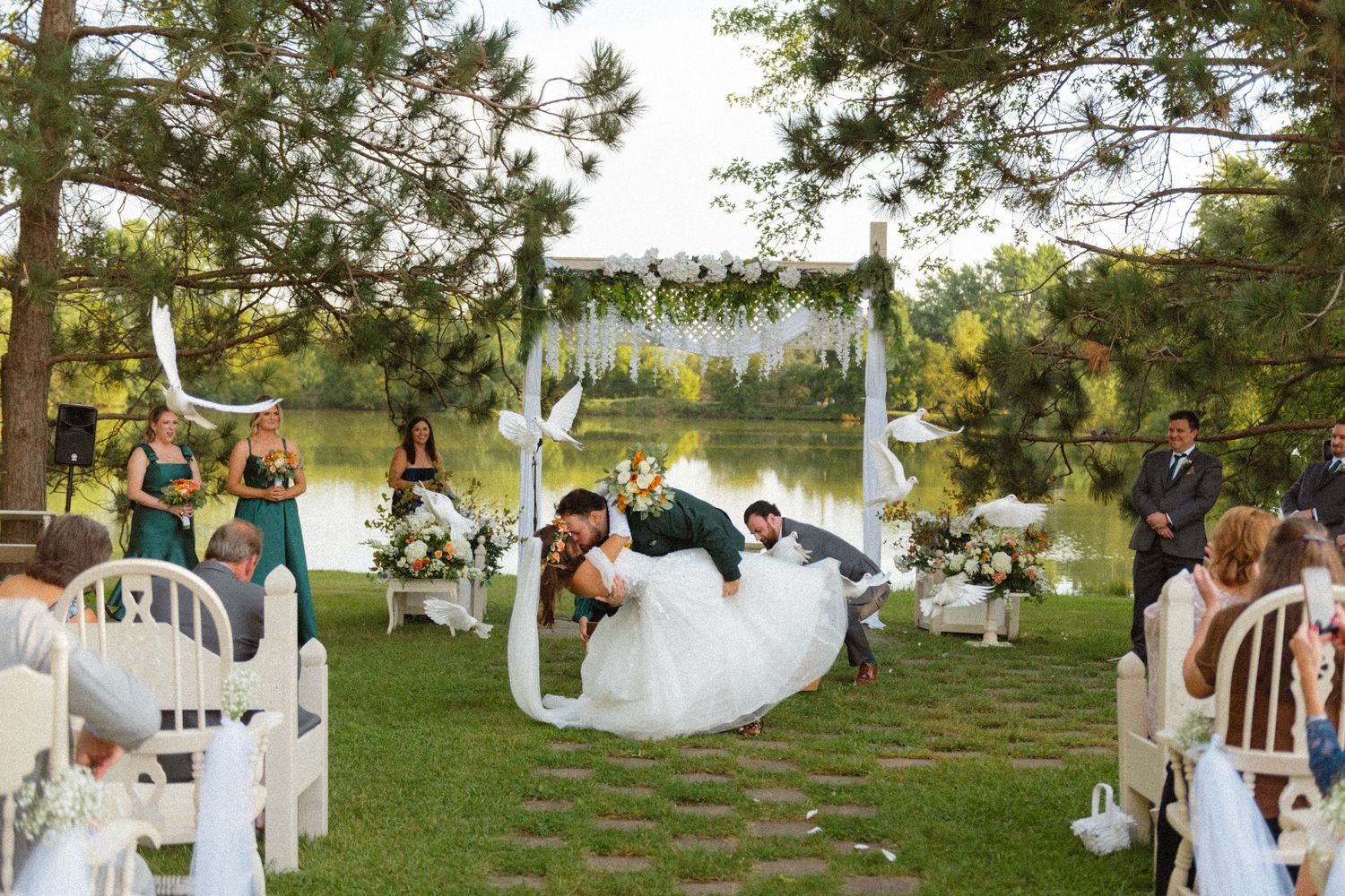 Bride and groom sharing romantic dip kiss at outdoor wedding ceremony by lakeside arbor.