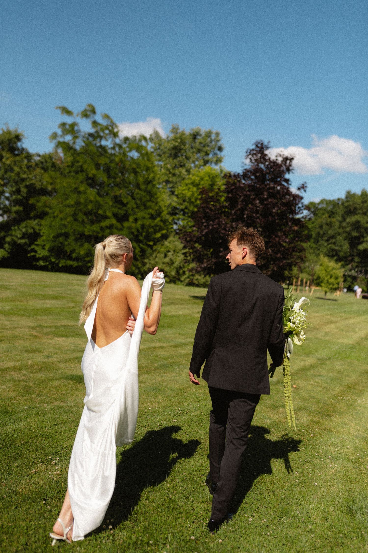 Bride and groom walk hand in hand across sunny green lawn.