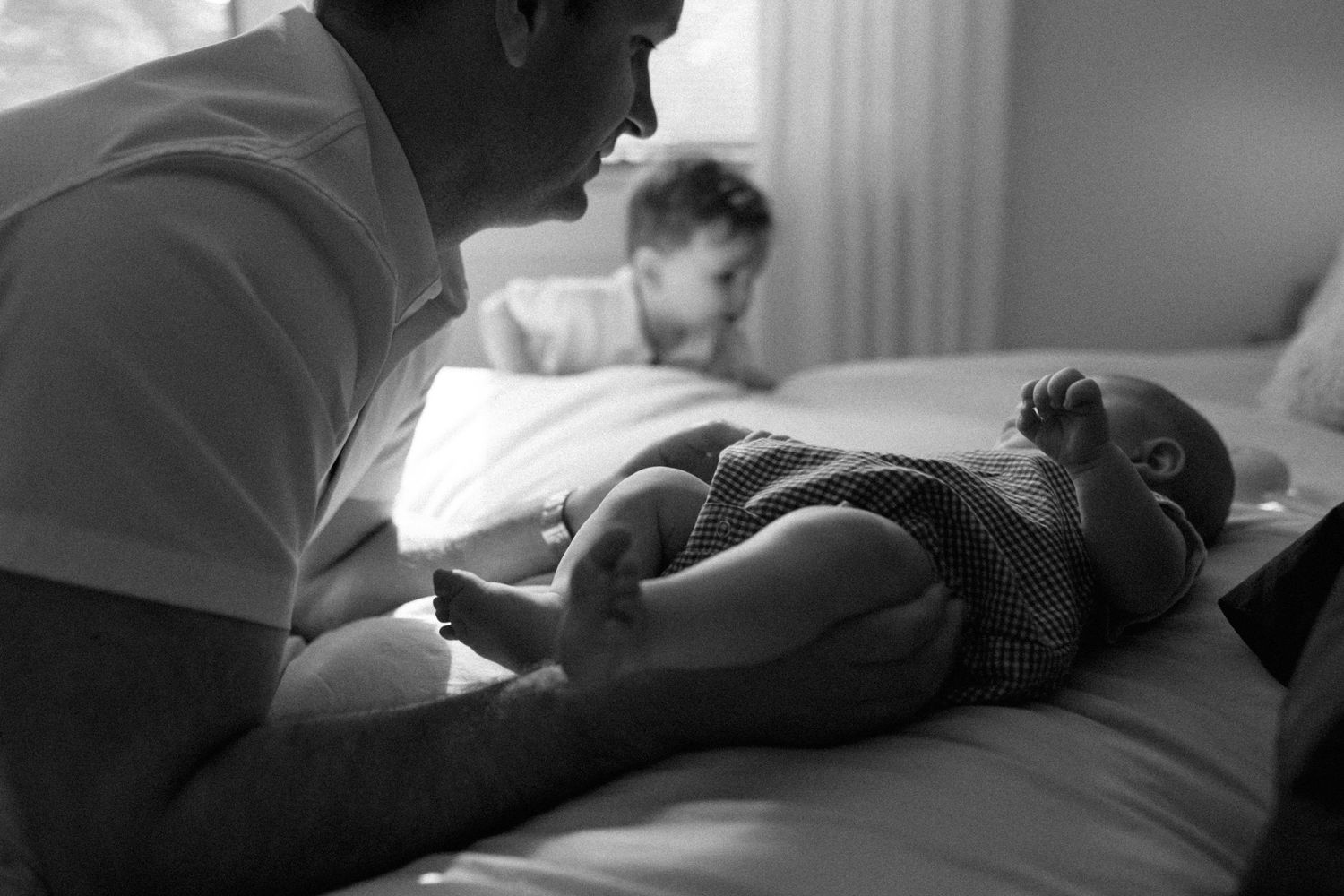 Black and white photo of father bonding with newborn on bed while toddler plays in background.