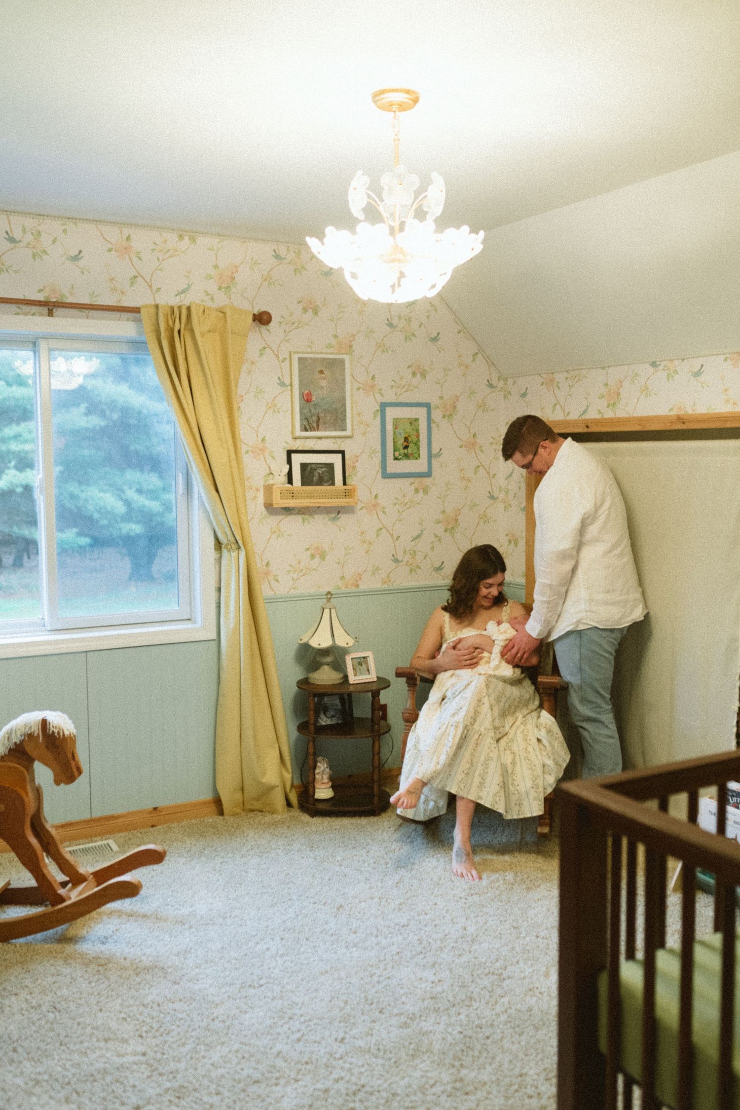 Parents with newborn baby in vintage nursery with floral wallpaper and rocking horse.