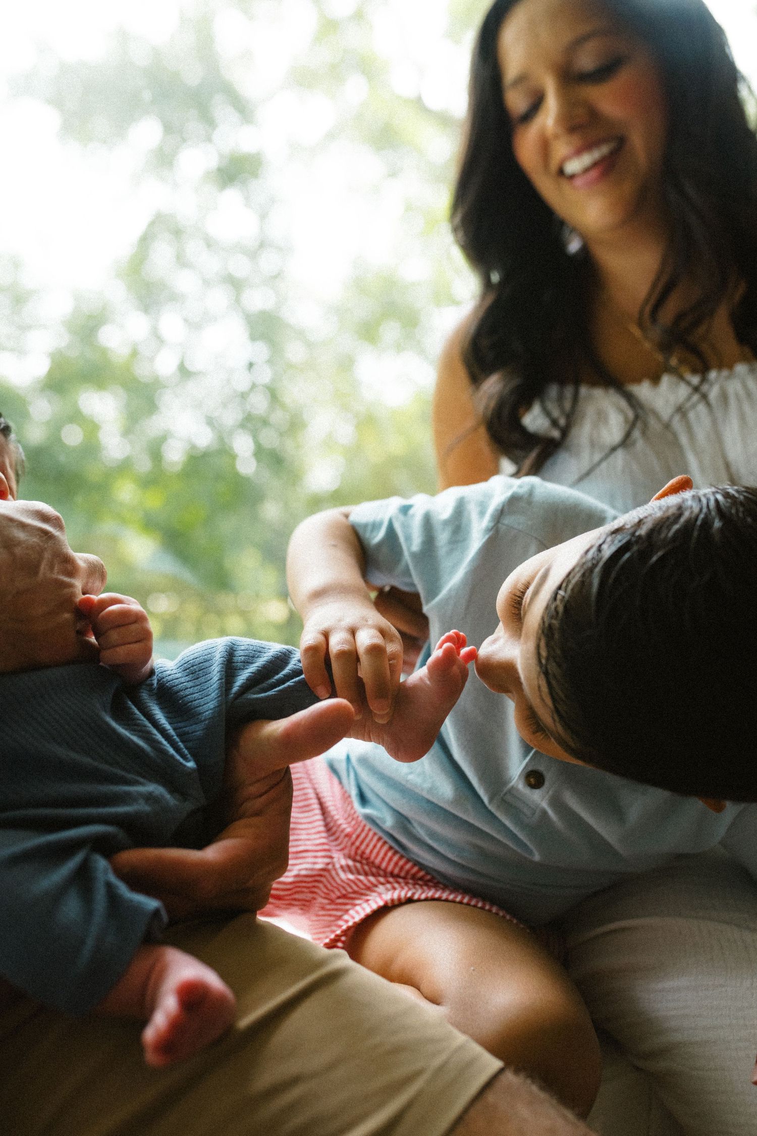 Smiling mother watches as young child touches newborn baby's tiny feet indoors near window.