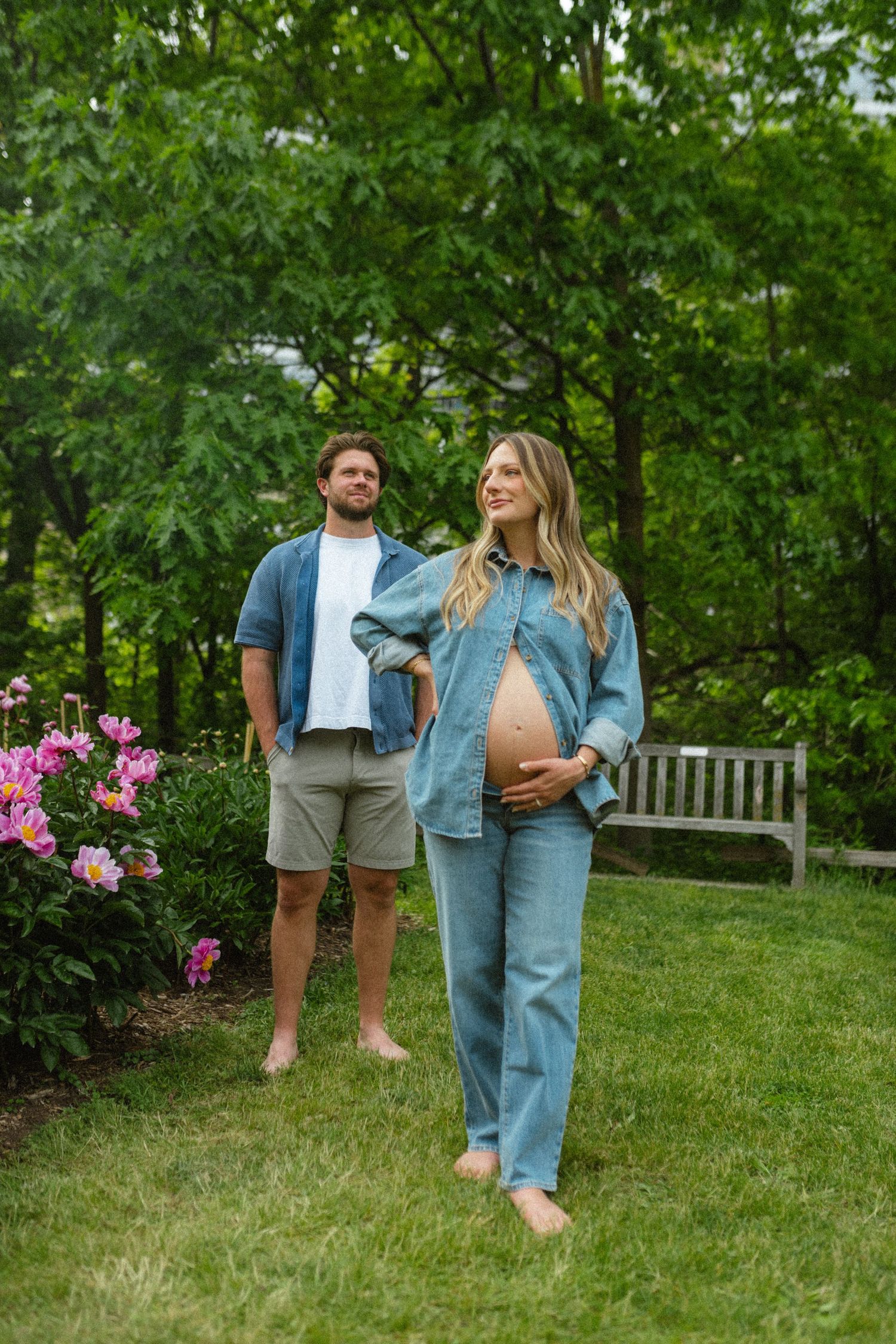 Pregnant woman in denim outfit cradling baby bump barefoot in lush green garden.